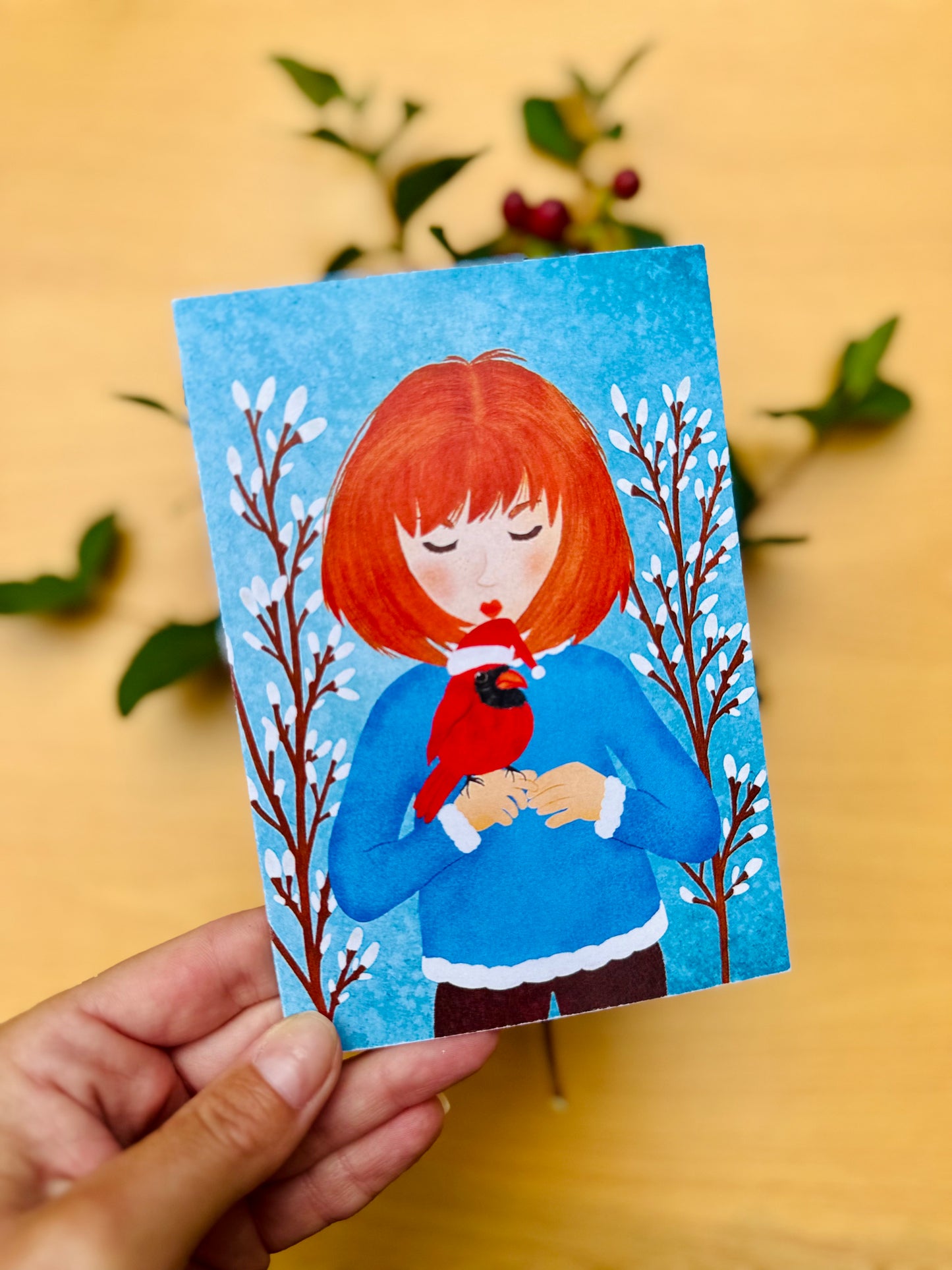 Hand holding a small card with a illustration of a person with red hair holding a bird, against a blue background.