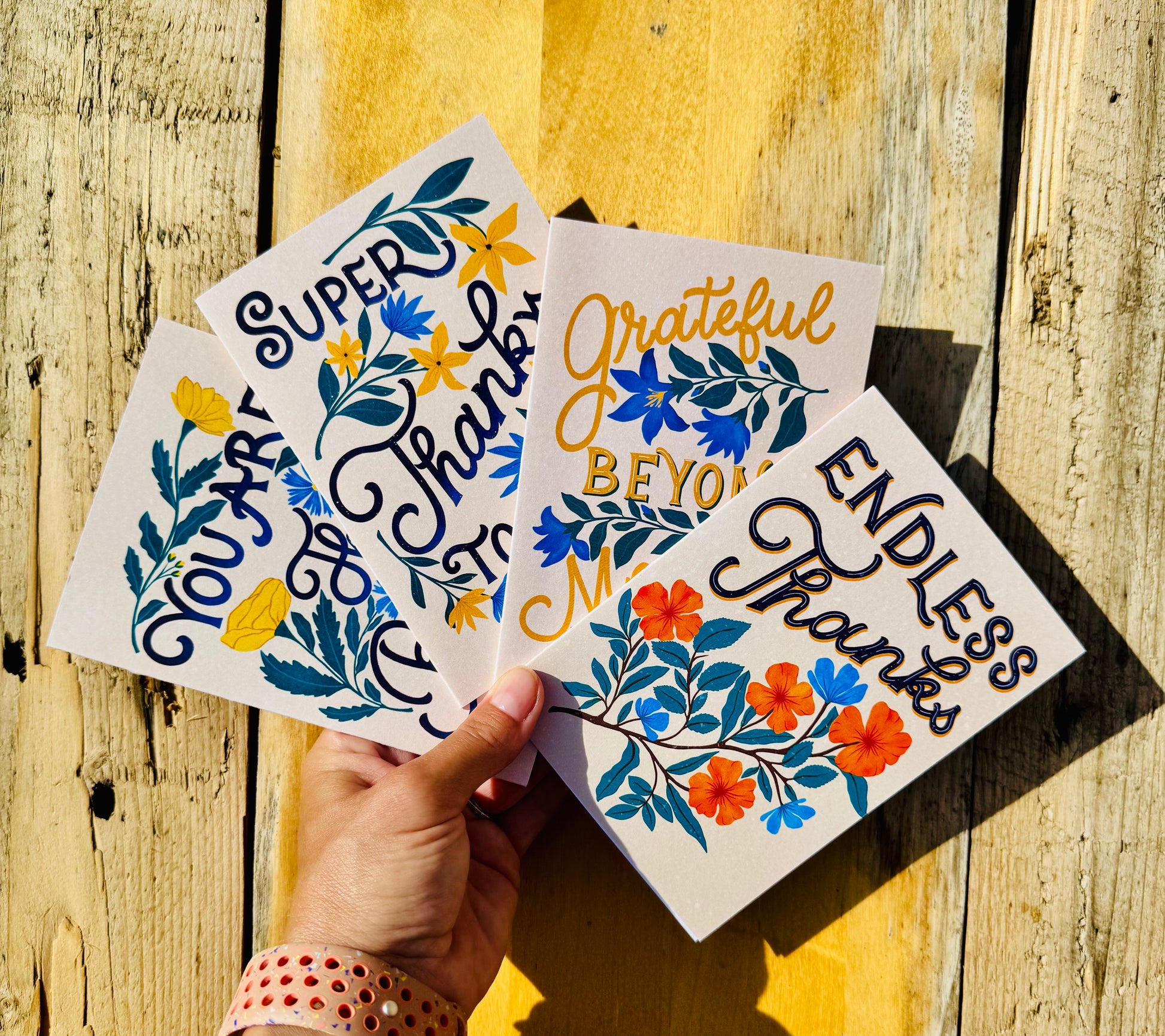 A hand holds four white art cards with colorful floral designs and inspirational hand-lettered text against a rustic wooden background. The cards read 'You Are Enough,' 'Super Grateful,' 'Grateful BEYOND Me,' and 'Endless Thanks.