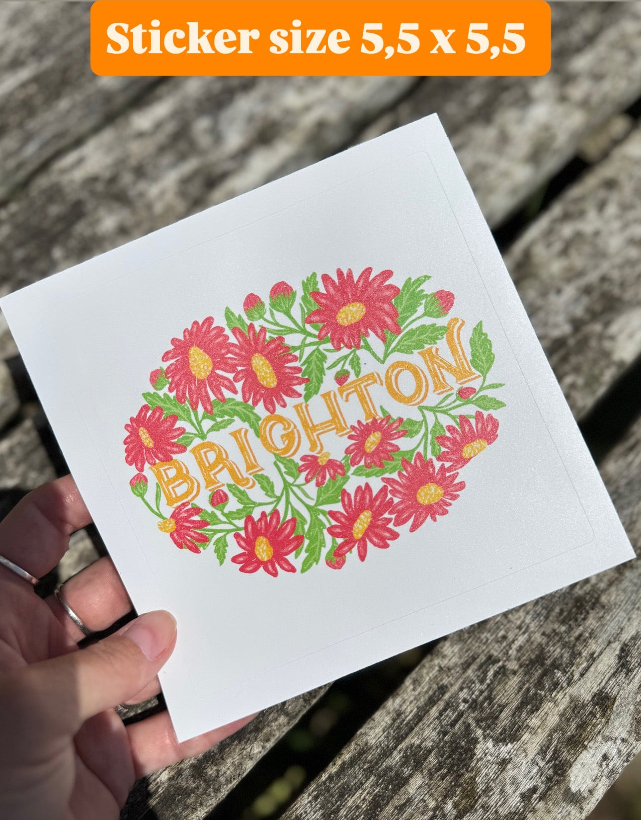 Hand holding a white card with floral design and 'Brighton' text, placed on a wooden surface.