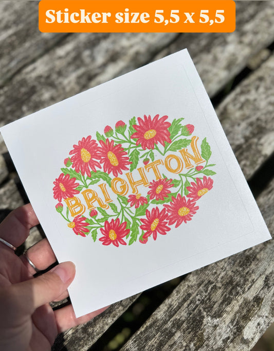 Hand holding a white card with floral design and 'Brighton' text, placed on a wooden surface.