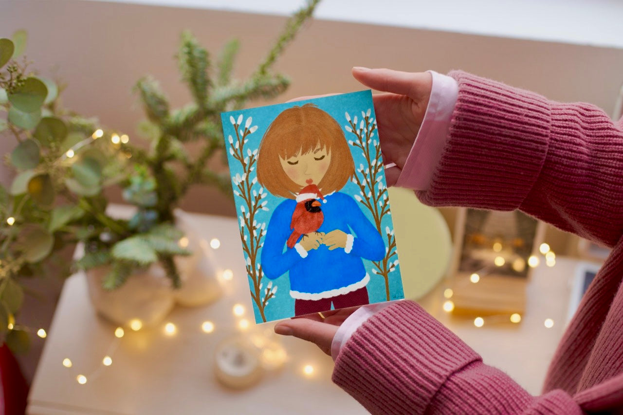 Person holding a card with a illustration of a person holding a robin, surrounded by plants and lights.