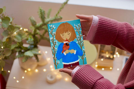 Person holding a card with a illustration of a person holding a robin, surrounded by plants and lights.