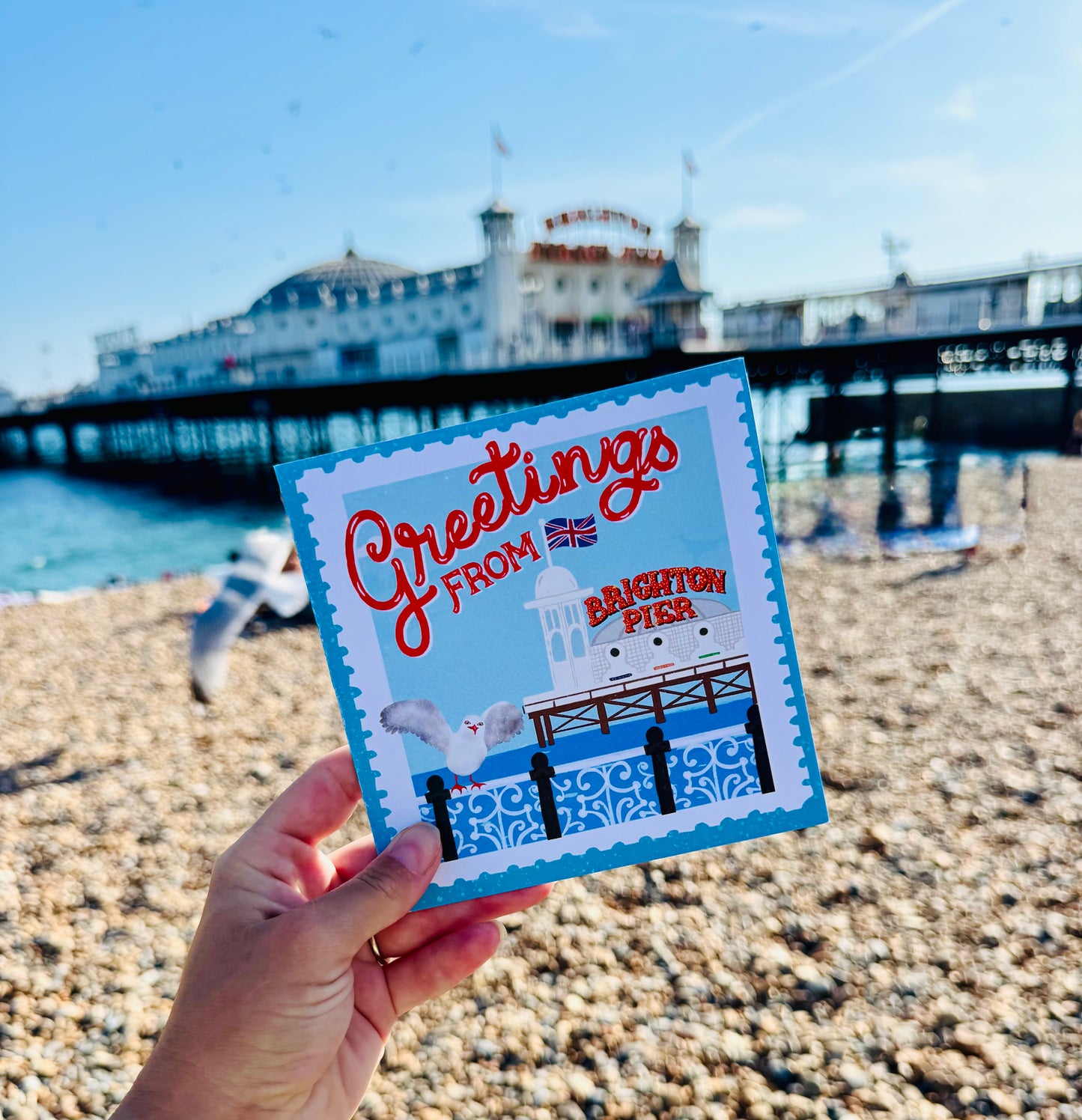 Hand holding a 'Greetings from Brighton Pier' card with a pier in the background on a beach.