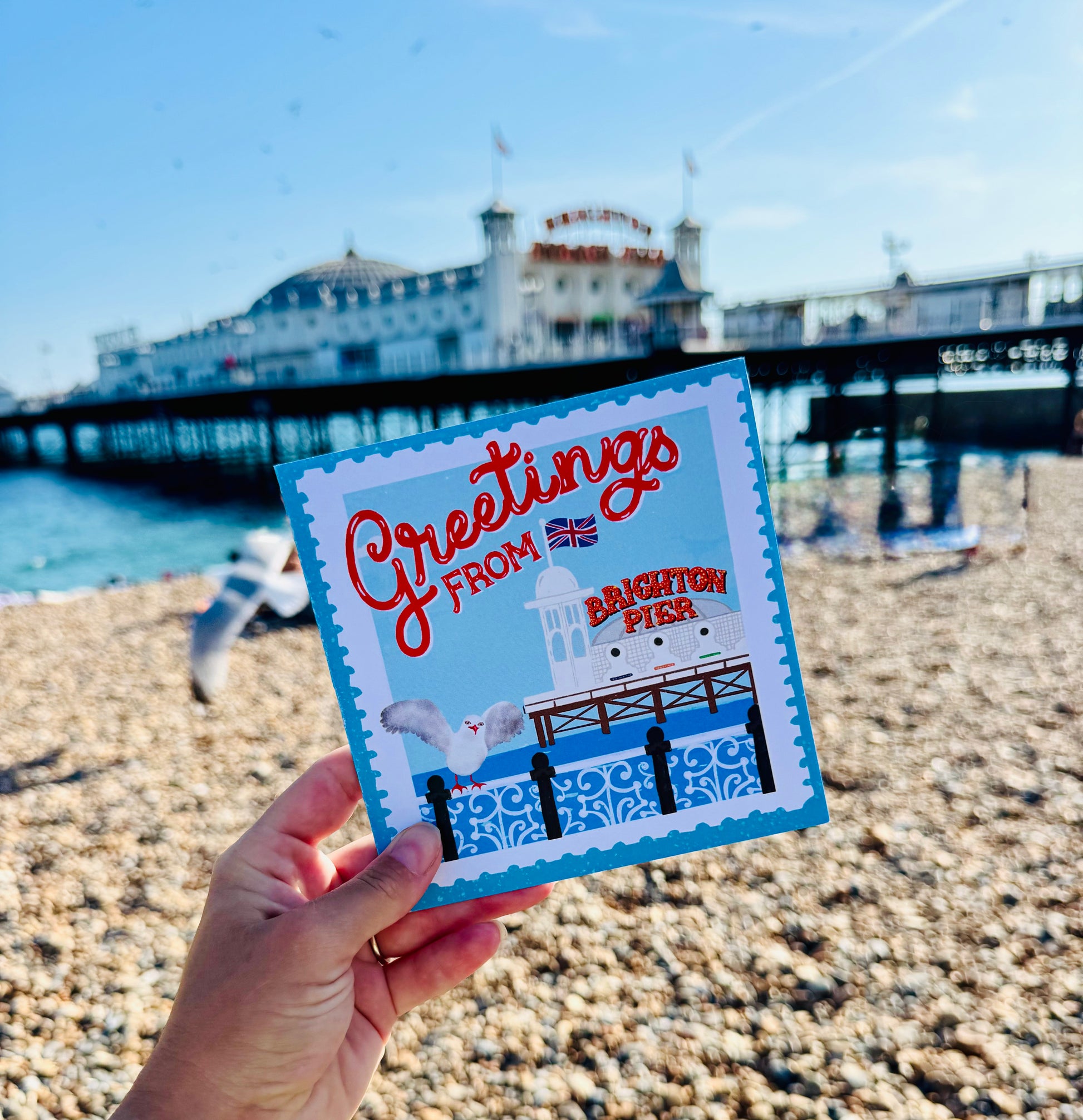 Hand holding a 'Greetings from Brighton Pier' card with a pier in the background on a beach.