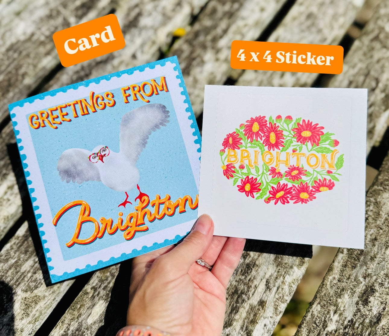 Hand holding a card and a sticker with 'Greetings from Brighton' text on a wooden surface.