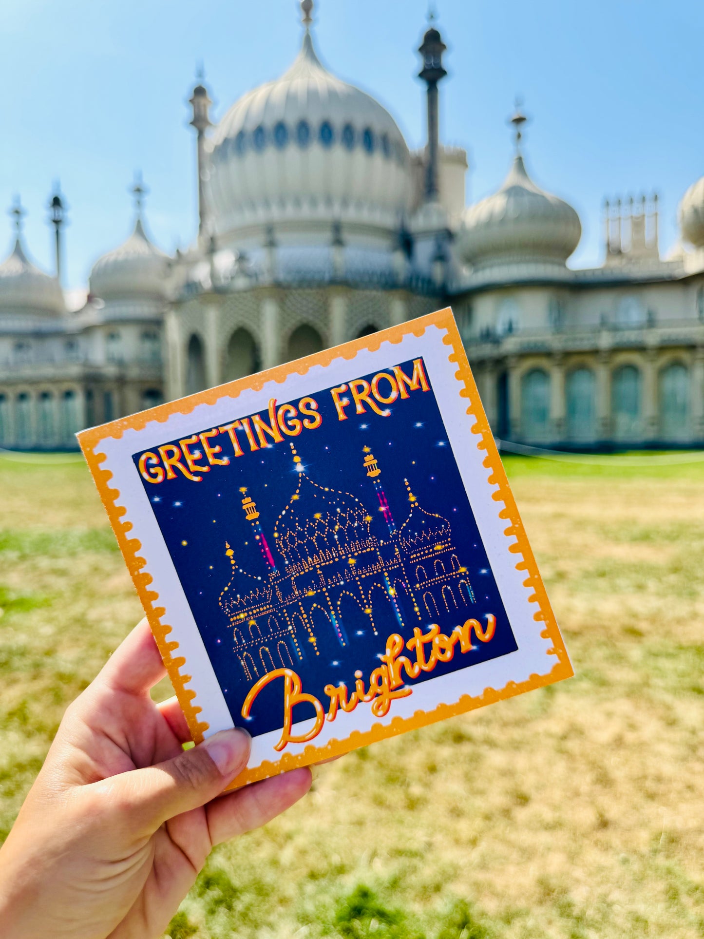 Card with 'Greetings from Brighton' text held in front of a royal Pavilion in Brighton building with domes.