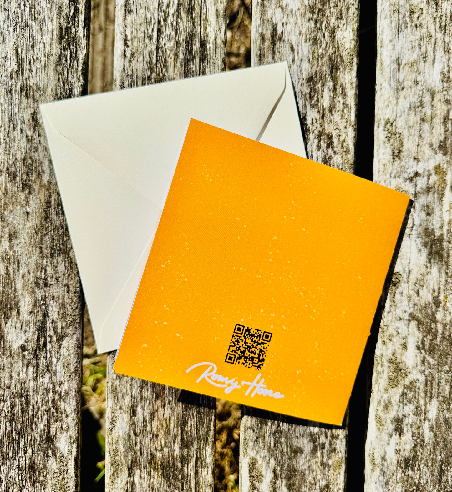 A square orange greeting card with a white envelope, placed on a weathered wooden surface. The card has a small QR code and the handwritten text 'Ready Home' in white script near the bottom.