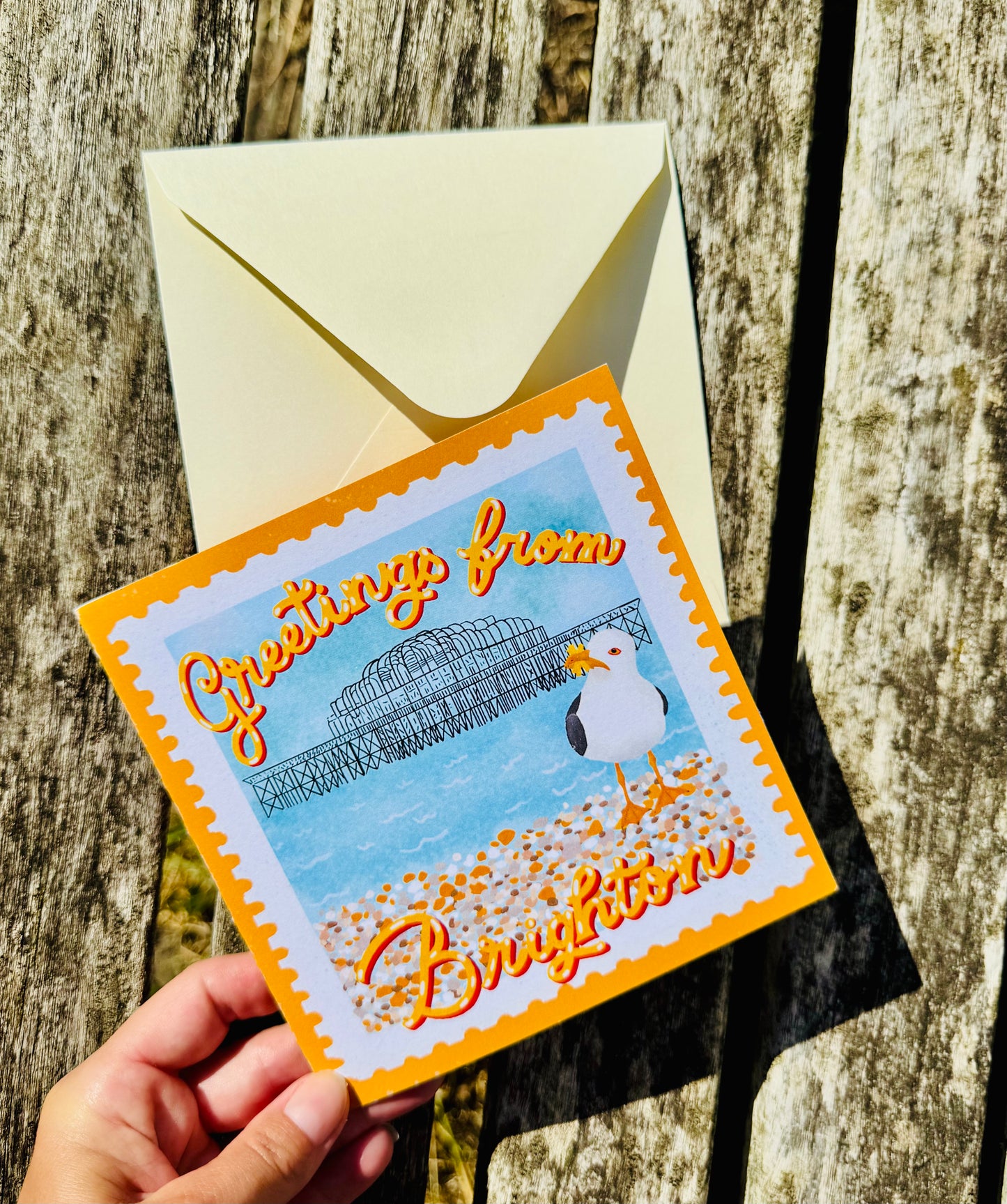 A hand holds a square greeting card with a bright orange border and an illustrated seagull on a beach, with the words 'Greetings from Brighton.' A cream envelope sits behind the card on a weathered wooden surface.