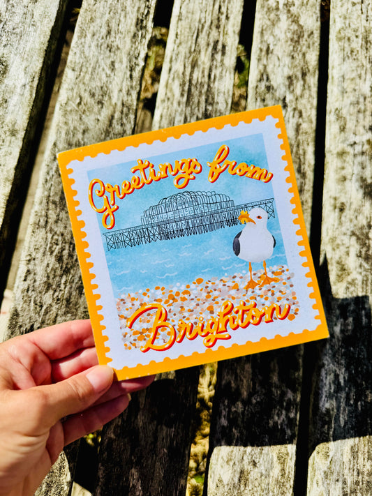A hand holds a square Brighton greeting card outdoors against a weathered wooden plank background. The card features a scalloped orange border, a blue sea-and-old pier in Brighton illustration, a white seagull, and orange script text reading 'Greetings from Brighton'.
