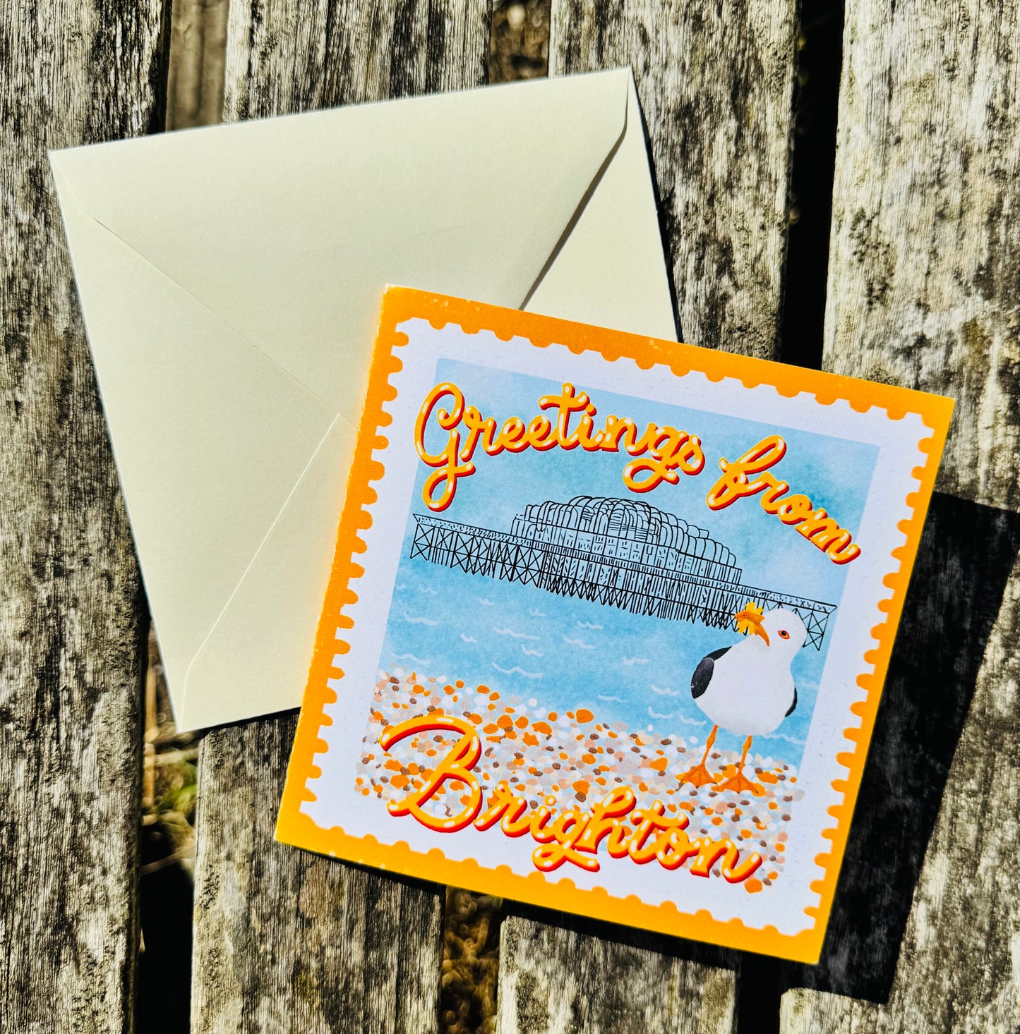 A square greeting card with a bright orange border and a seaside illustration reading 'Greetings from Brighton,' placed on a rustic weathered wood surface with a cream envelope behind it.