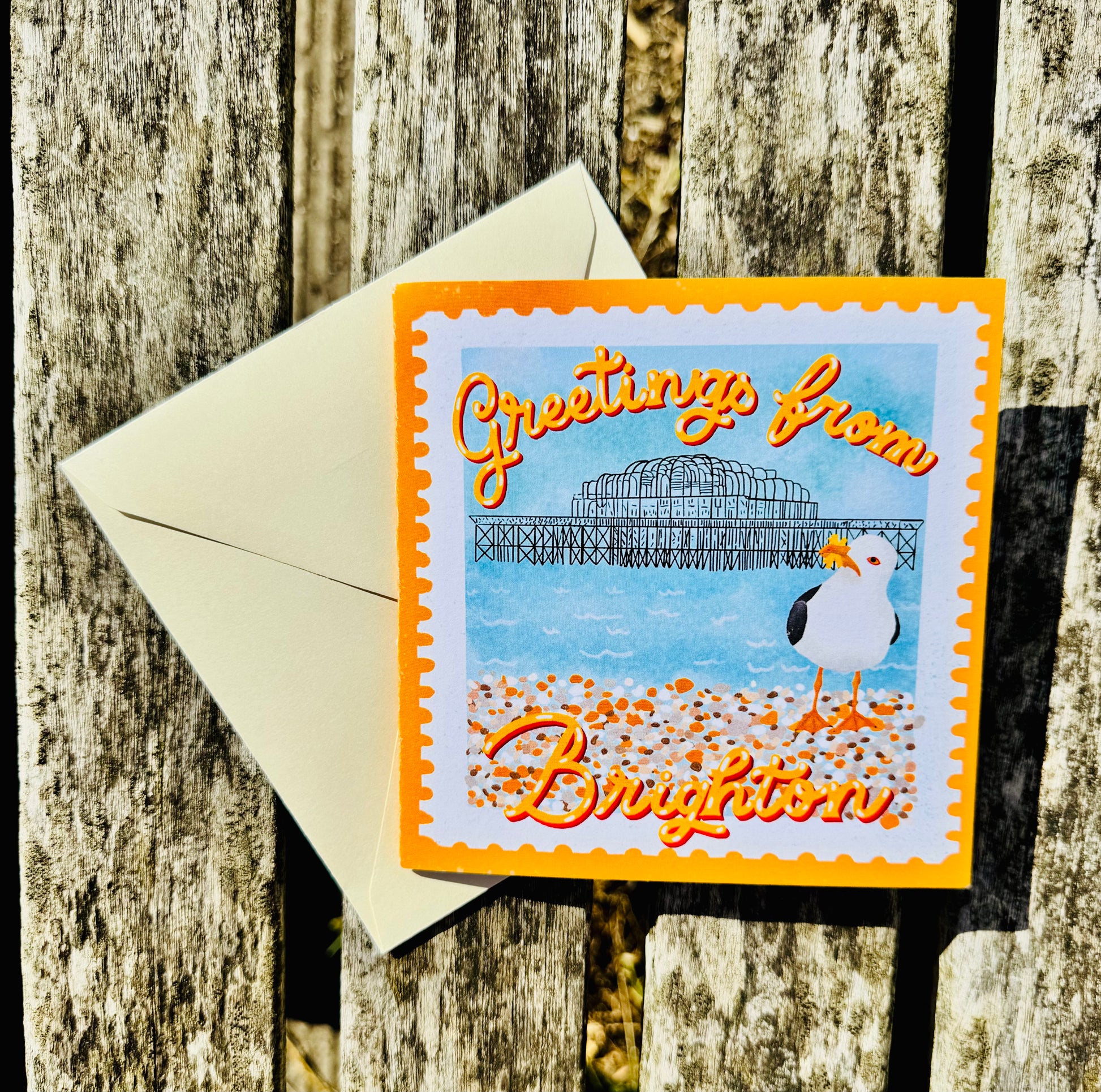 A square greeting card with a bright orange border and the words 'Greetings from Brighton' in orange script, placed on top of an off-white envelope against a weathered wooden surface. The card features a beach scene with a pier, blue sky, and a seagull.