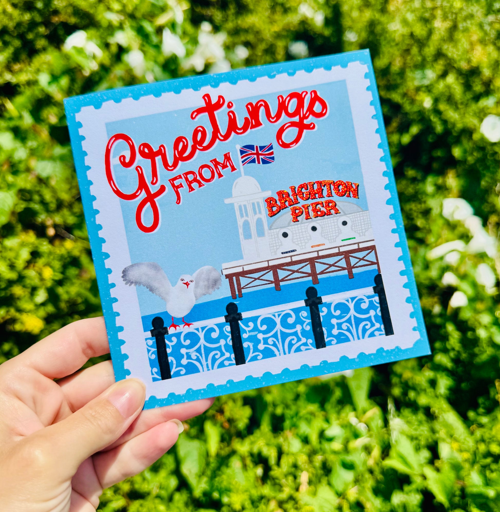 Greeting card with 'Greetings from Brighton Pier' text and illustration held in front of green foliage.
