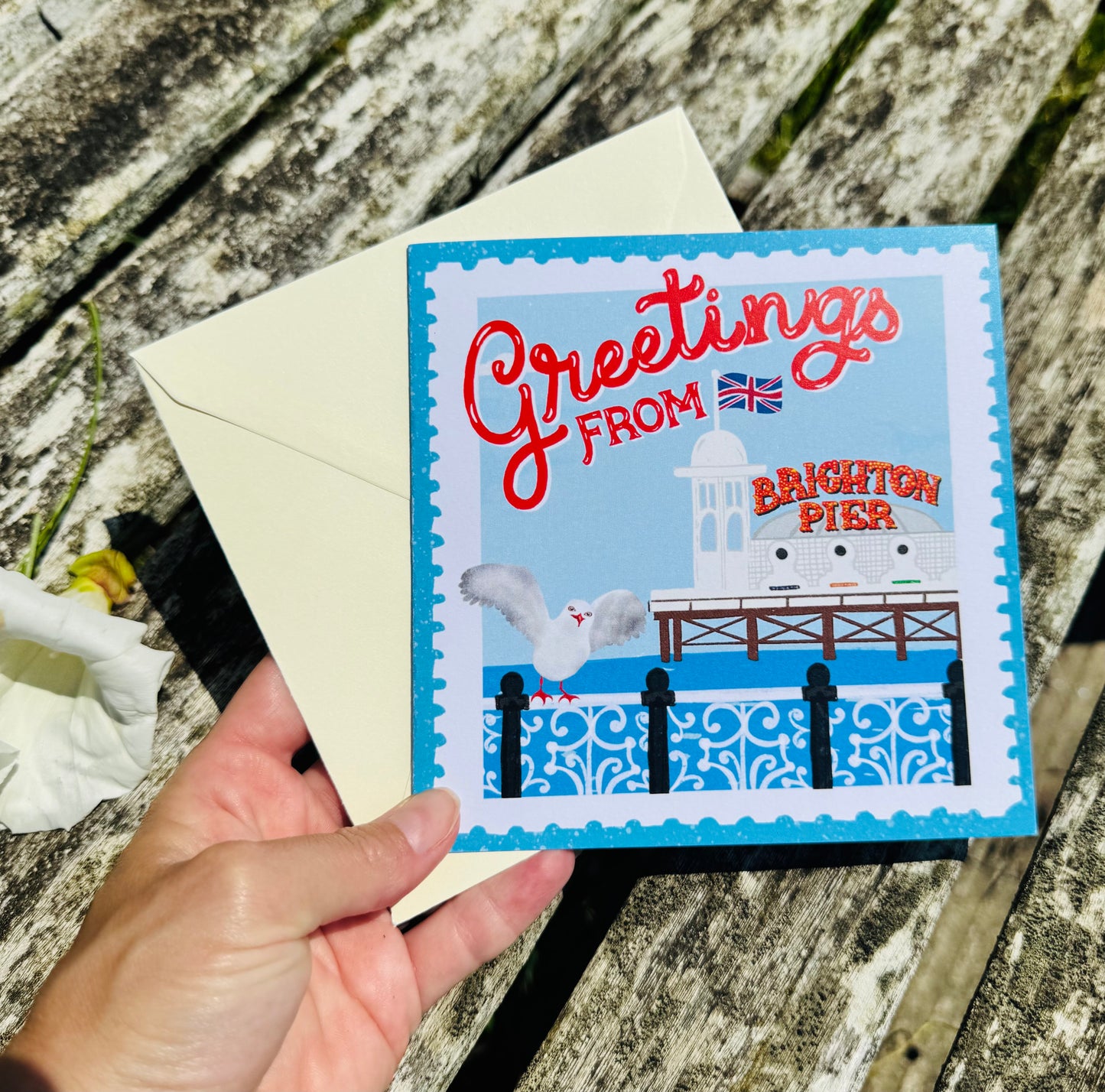 Greeting card with 'Greetings from Brighton Pier' text and illustration, held over a wooden surface.