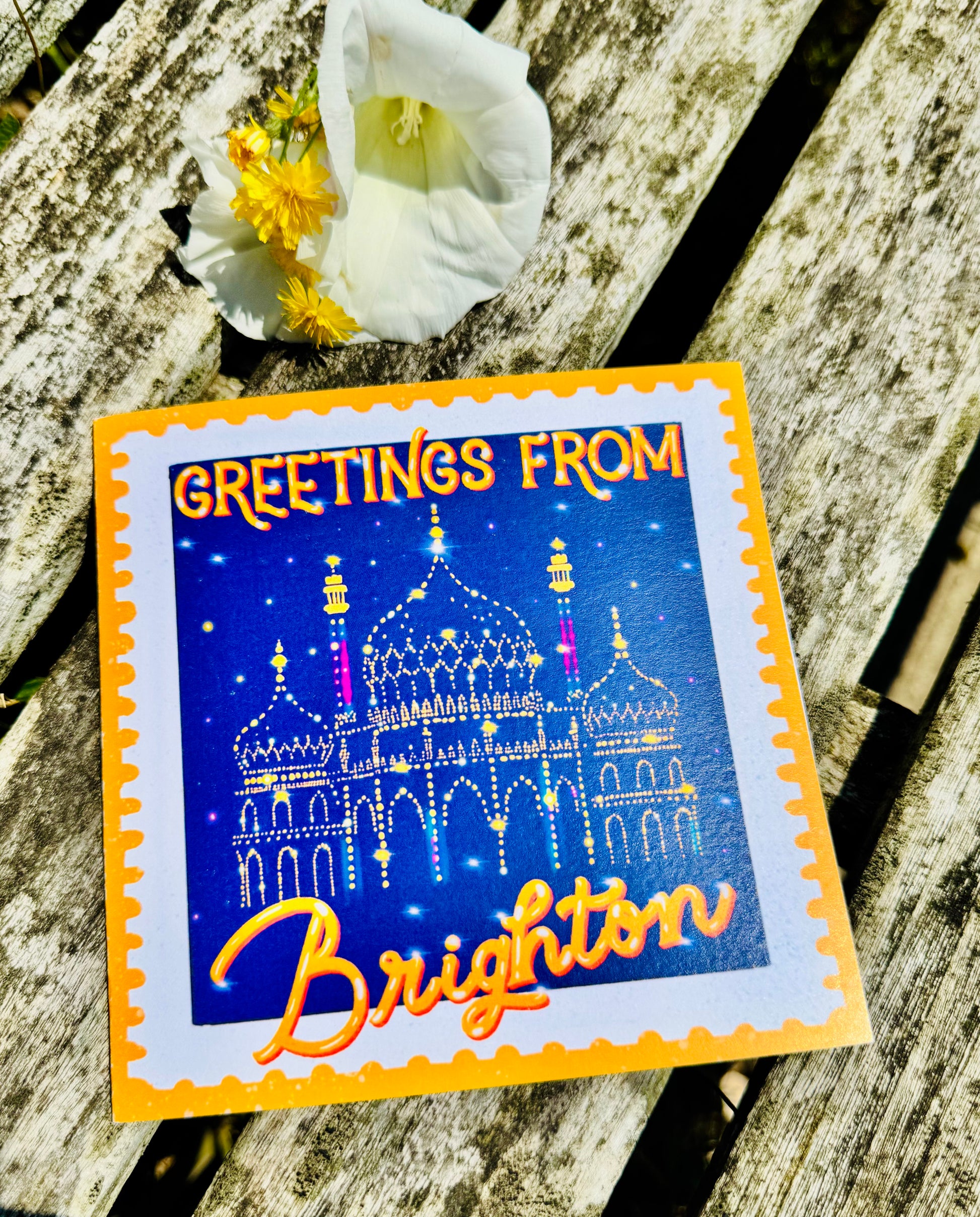 Greeting card with 'Greetings from Brighton' text and a decorative design on a wooden surface with flowers.