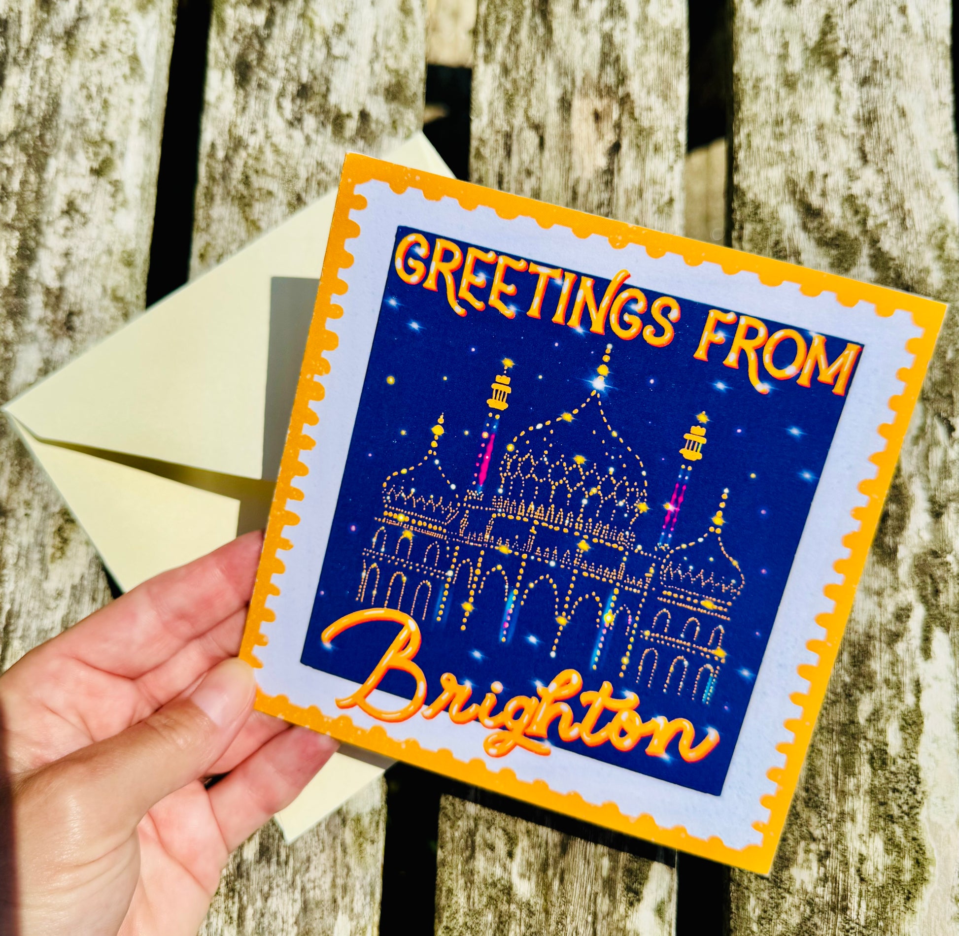 Greeting card with 'Greetings from Brighton' text and a castle illustration held by a hand against a wooden background.