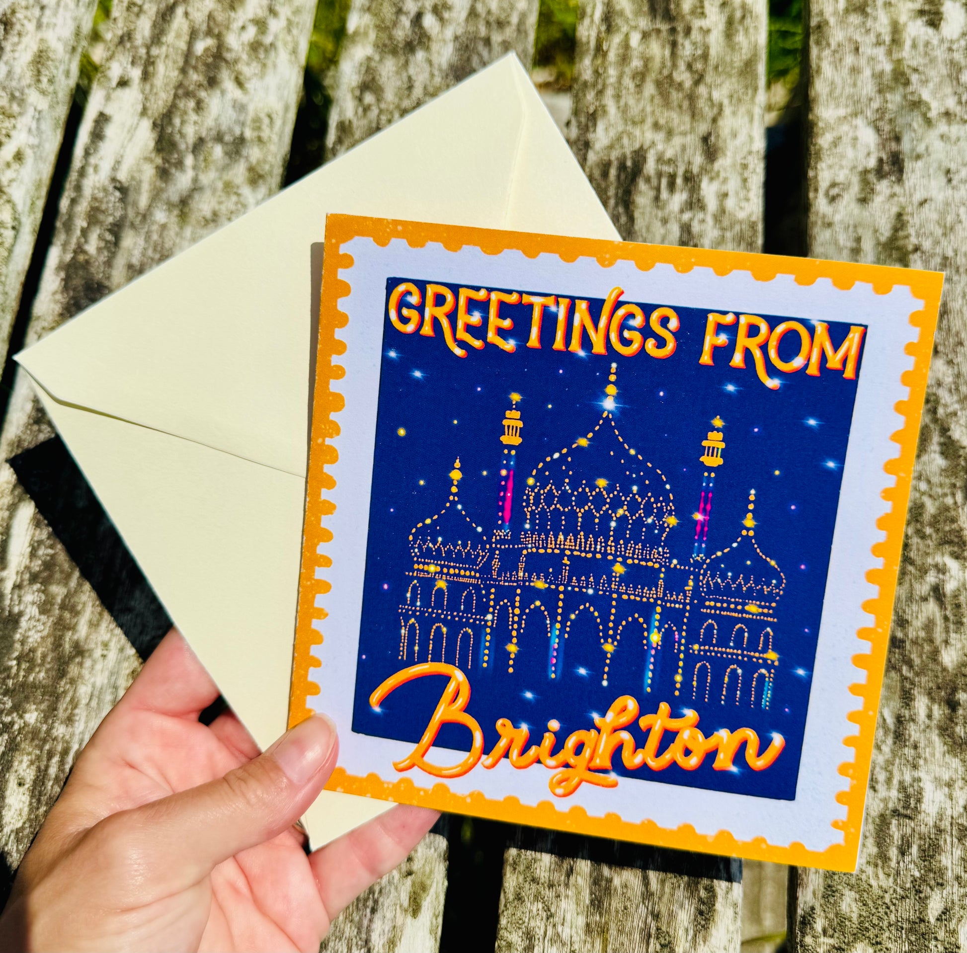 Greeting card with 'Greetings from Brighton' text and a building illustration, held over an envelope against a wooden background.