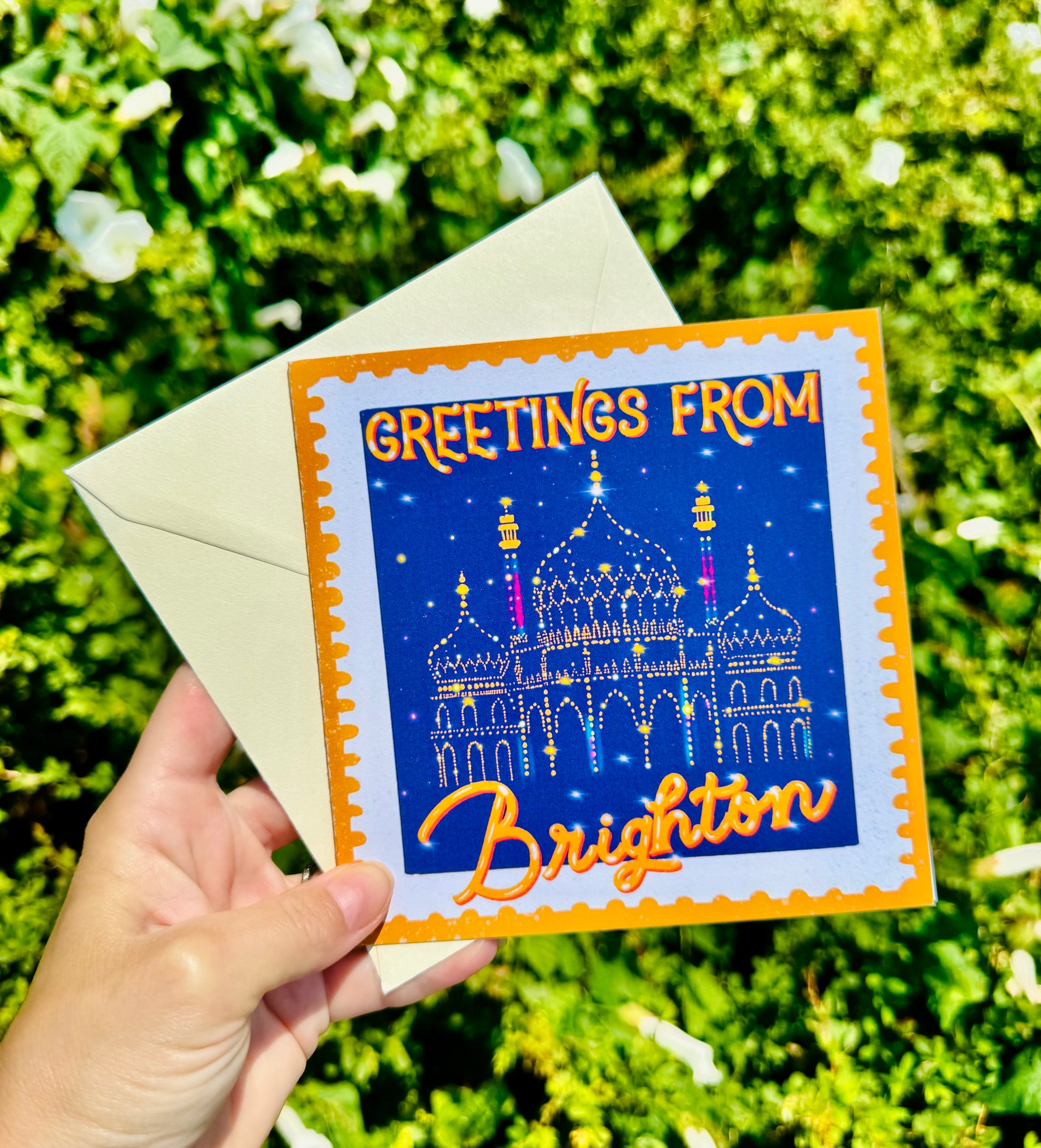 Greeting card with 'Greetings from Brighton' text held against a green leafy background