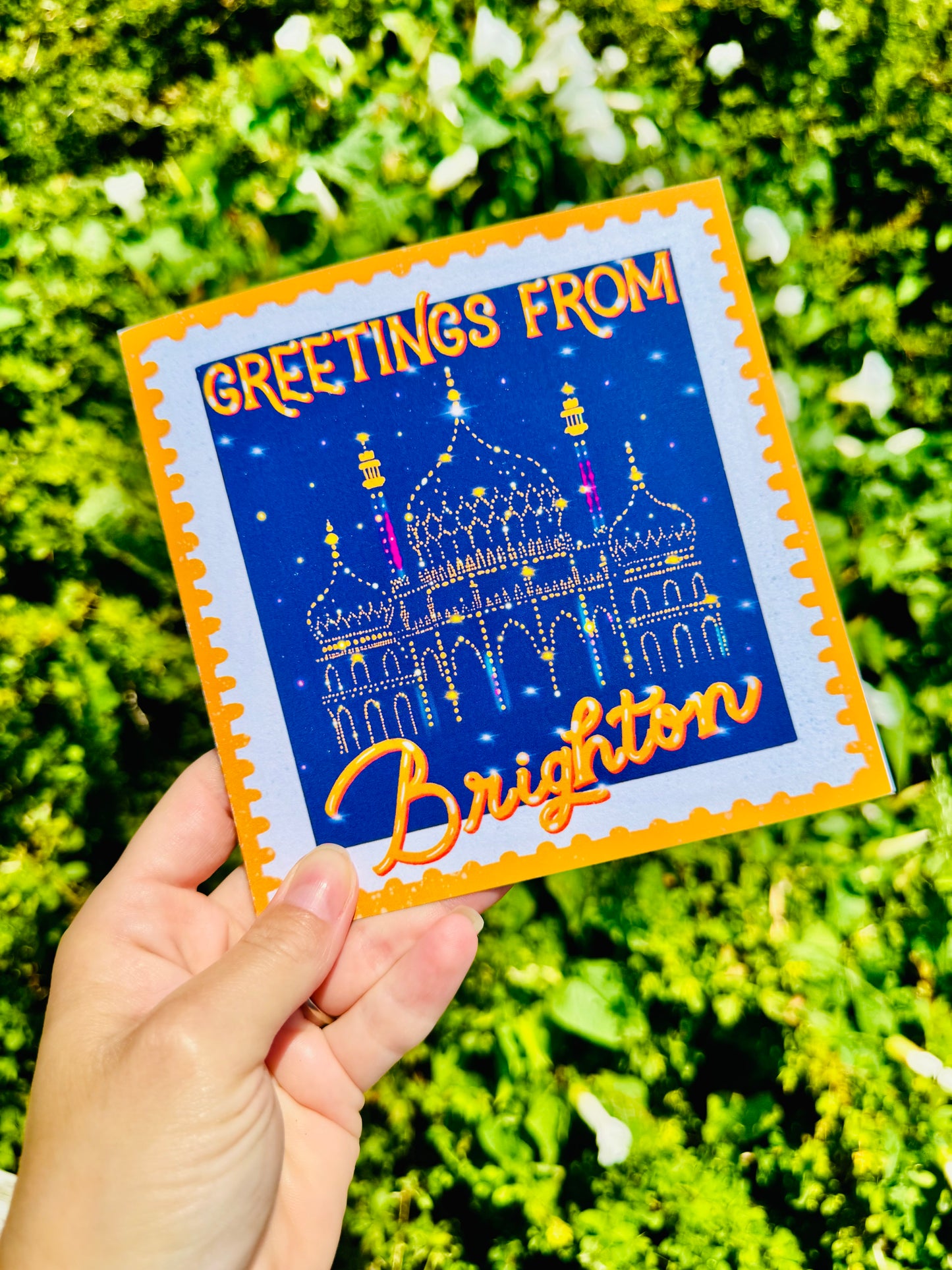 Hand holding a postcard with 'Greetings from Brighton' text and a castle illustration against a green leafy background.