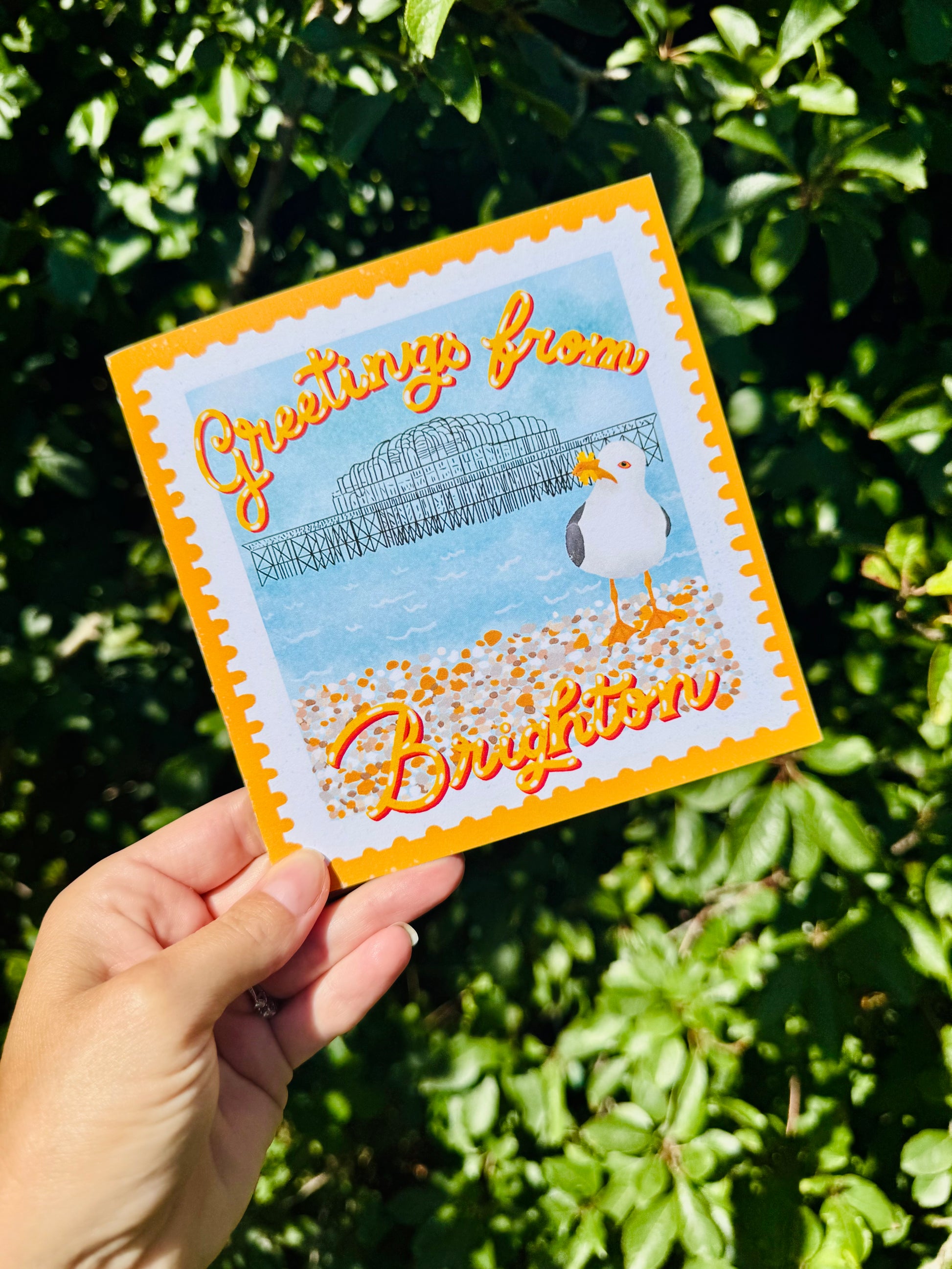 A hand holds a square greeting card that reads 'Greetings from Brighton' with a seagull and Brighton Pier illustration, set against a background of green foliage.
