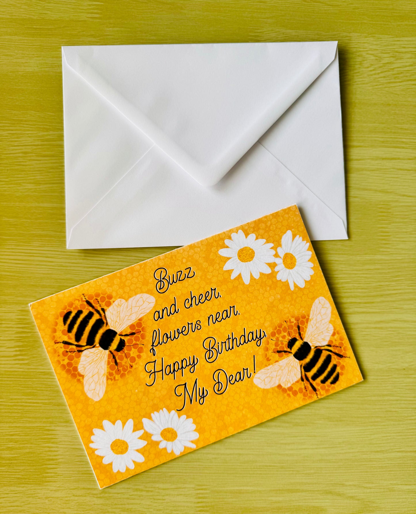 Birthday card with bees and flowers on an orange background, accompanied by a white envelope on a green surface.