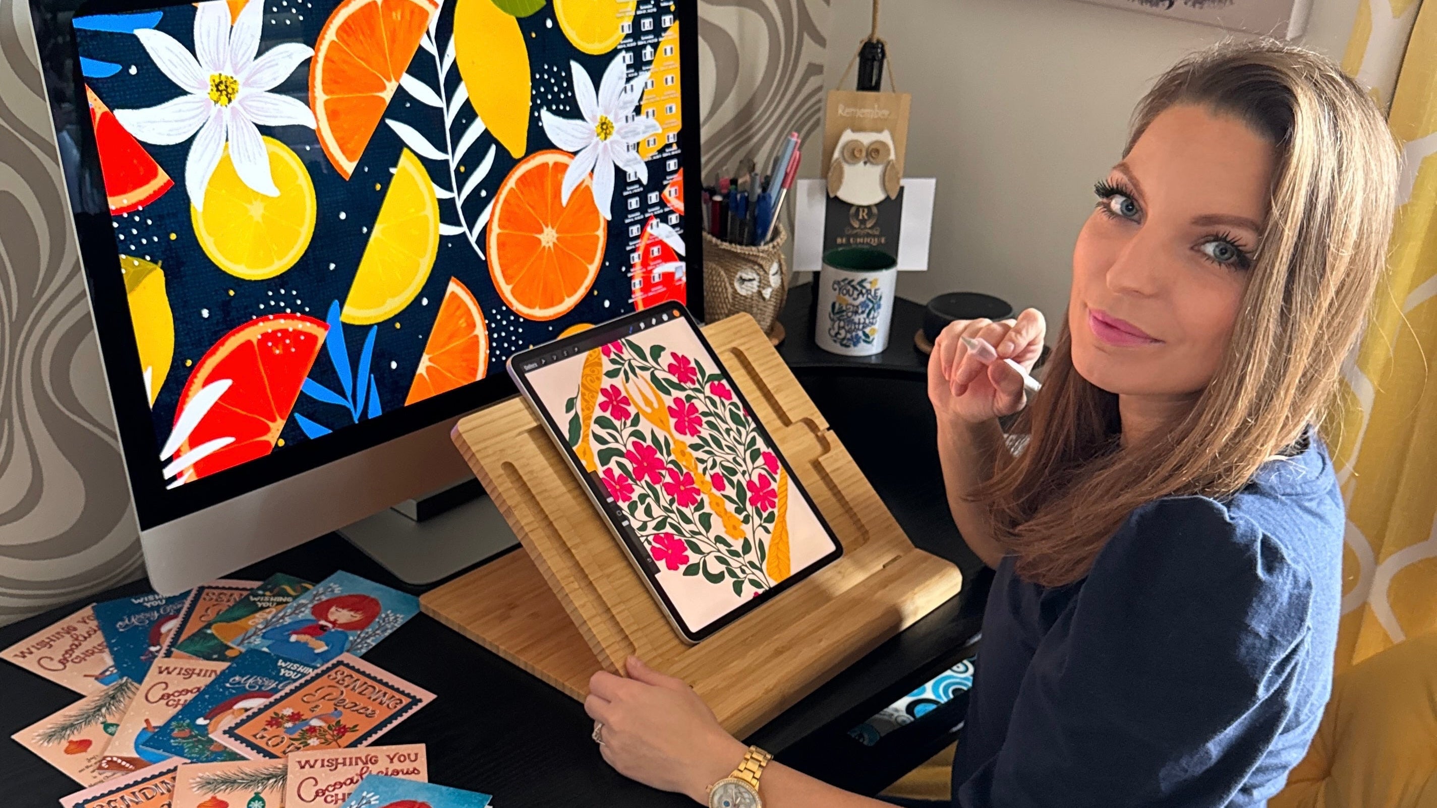 Woman sitting at a desk with a computer displaying colorful artwork, surrounded by art supplies.