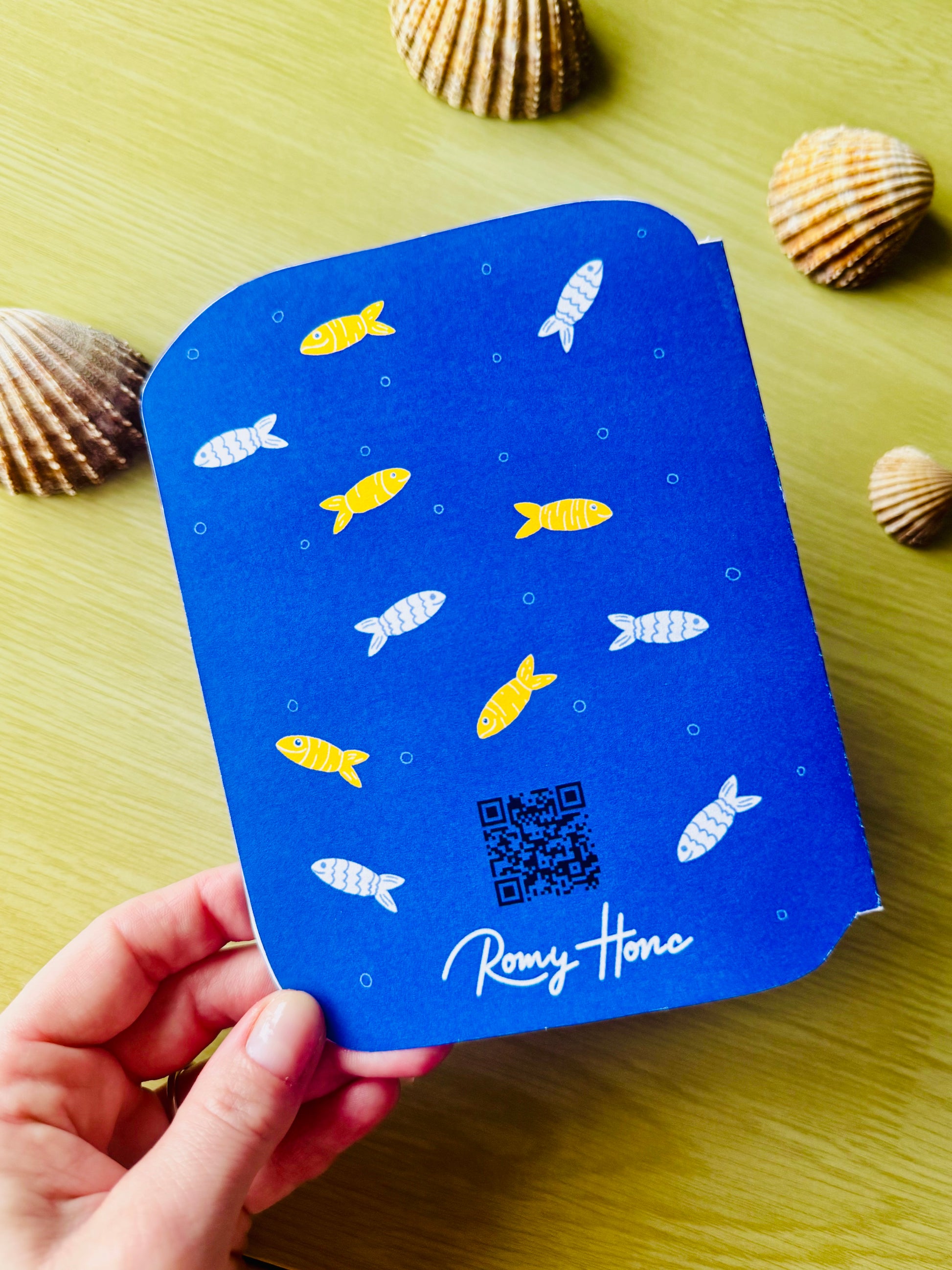 Back side of the Blue birthday card with fish patterns and a QR code held by a hand on a wooden surface with shells.
