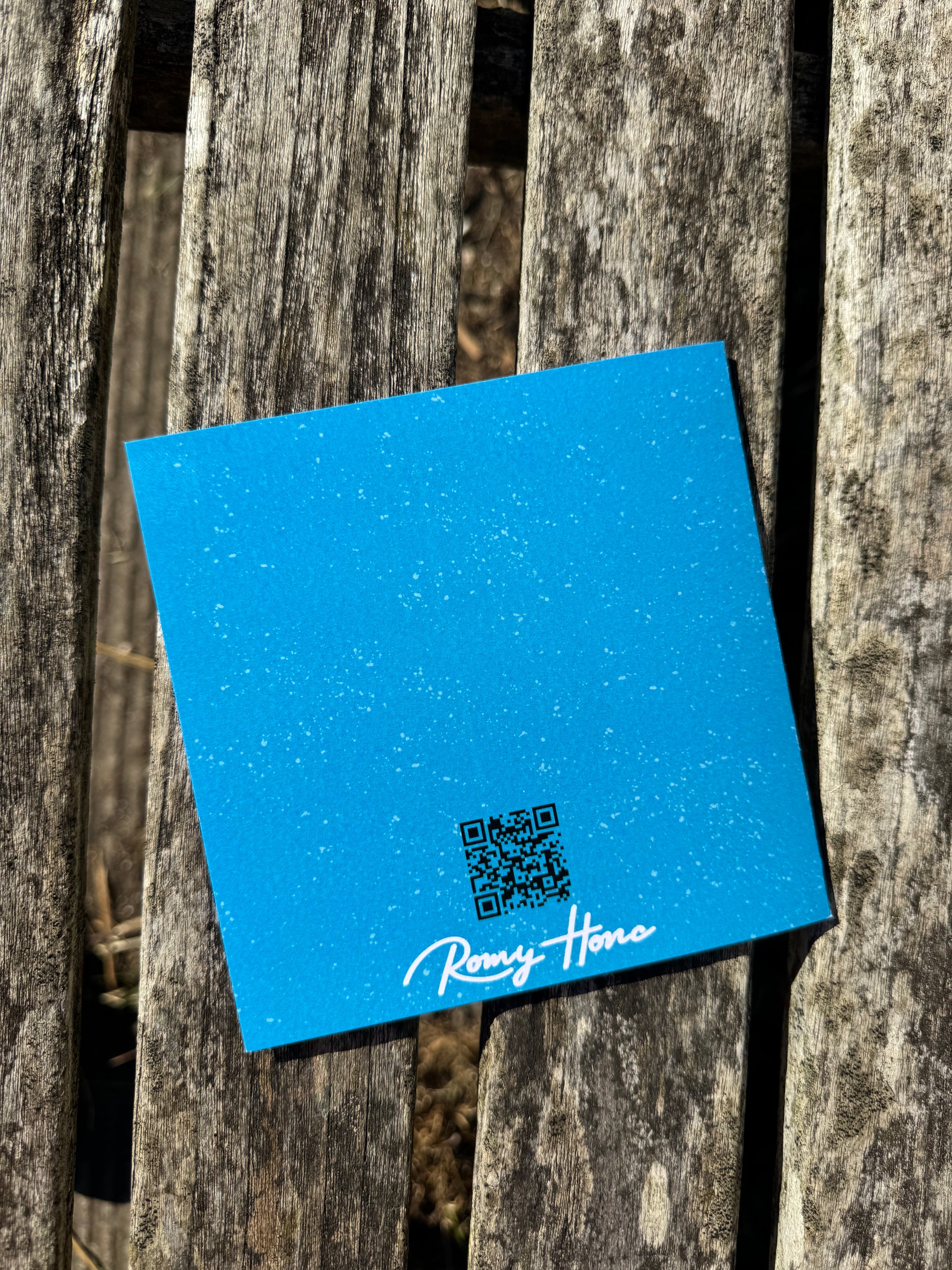 A square blue card or sticker with a small QR code and the white script text 'Romy Honc' lies on weathered wooden boards outdoors.