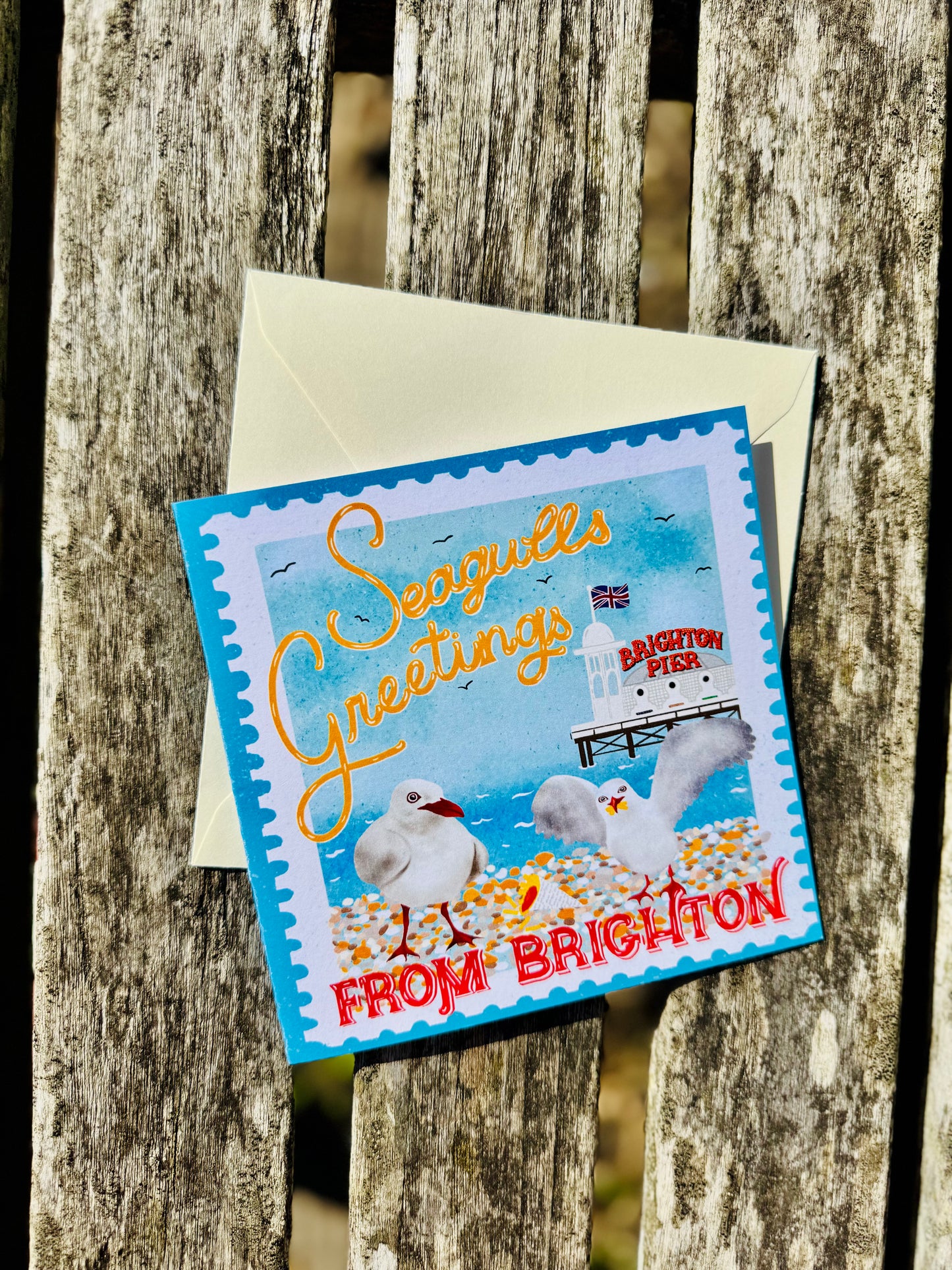 A square seagull-themed greeting card with a cream envelope, placed on a weathered wooden surface. The card reads 'Seagulls Greetings FROM BRIGHTON' and features illustrations of seagulls, a lighthouse, and the word 'Brighton' in red.