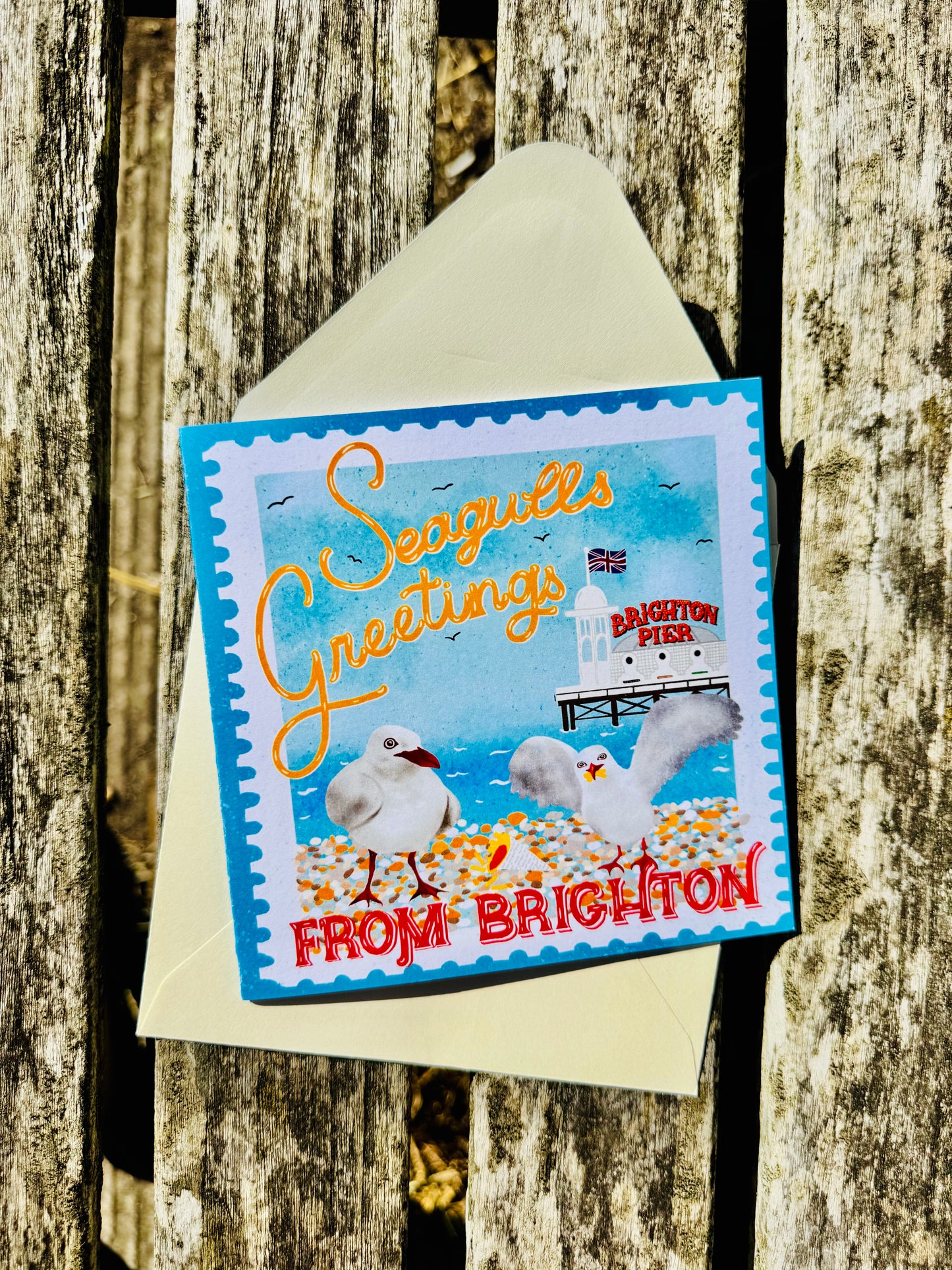 A square seagull-themed greeting card with a blue border and an illustrated lighthouse scene, placed on top of a cream envelope against a weathered wooden plank background. The card reads 'Seagulls Greetings' and 'FROM BRIGHTON.