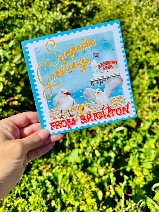 A hand holds a square postcard featuring a Brighton seagull illustration and the text 'Seagulls Greetings FROM BRIGHTON' against a blurred green outdoor background.