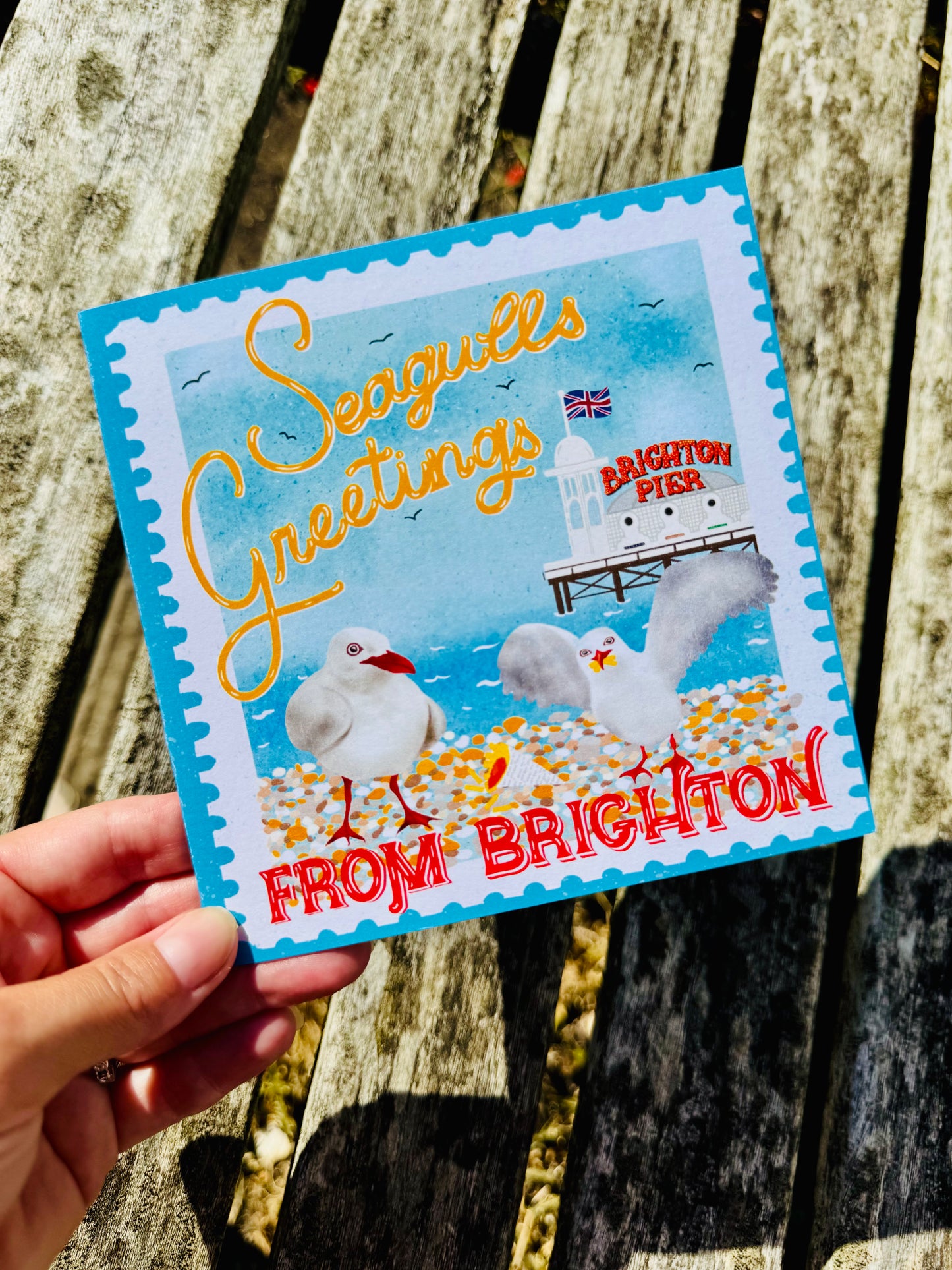 A hand holds a square Seagulls Greetings Brighton-themed card outdoors against a weathered wooden background. The card features a blue border, an illustration of seagulls, a lighthouse, and the text 'Seagulls Greetings' and 'FROM BRIGHTON'.
