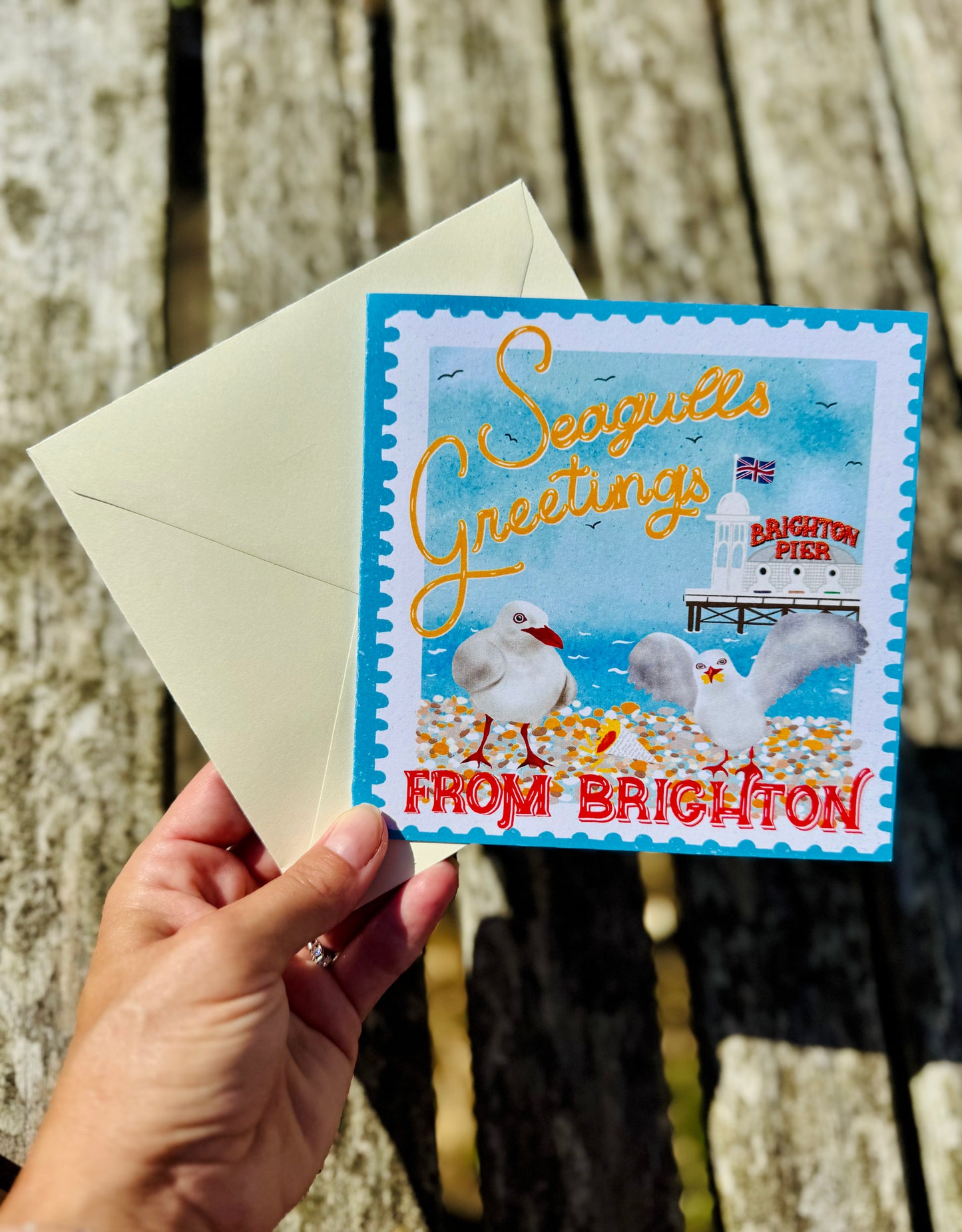 A hand holds a Brighton seagull greeting card with its cream envelope, set against a blurred wooden background. The card features a colorful illustration of seagulls, a pier, and the text 'Seagulls Greetings FROM BRIGHTON'.