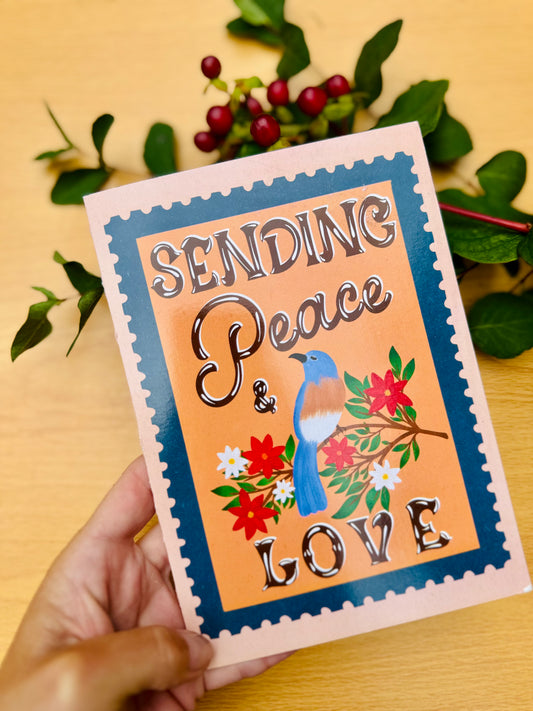 Card with 'Sending Peace & Love' text and a blue bird illustration, held by a hand on a wooden surface with leaves.
