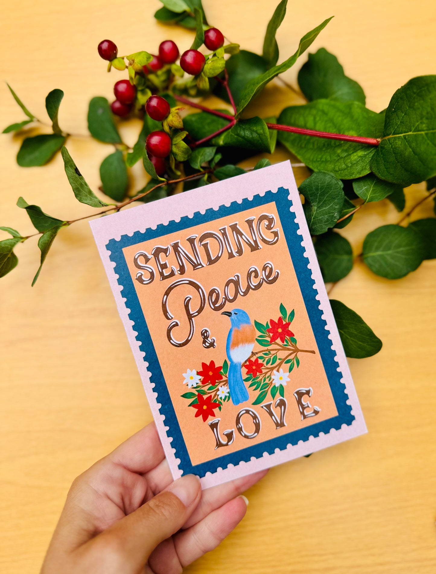 Hand holding a card with 'Sending Peace & Love' text and a bird illustration on a yellow background with green leaves.