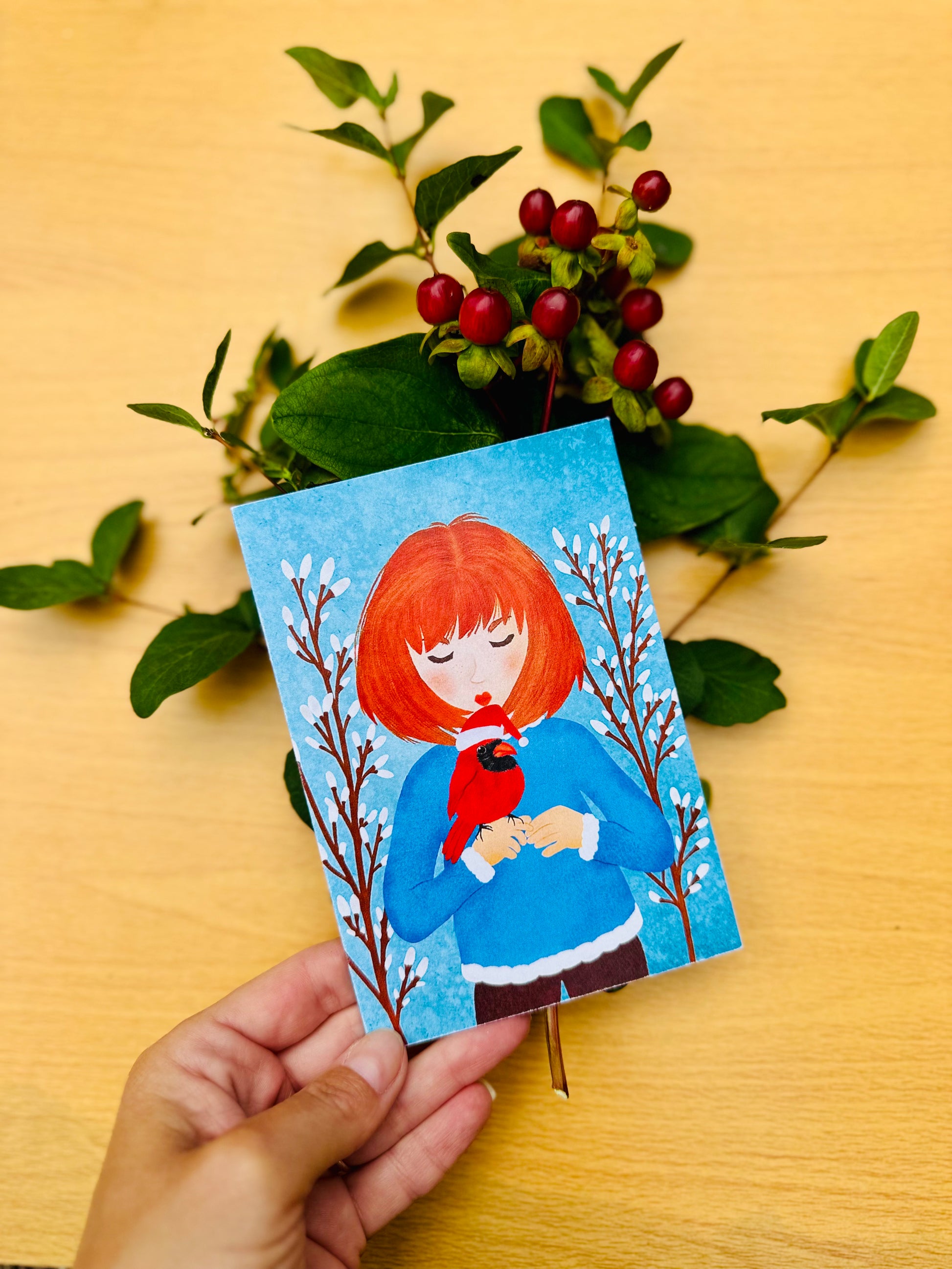 Hand holding a small card with a illustration of a person with red hair and a bird, surrounded by green leaves and berries on a wooden surface.