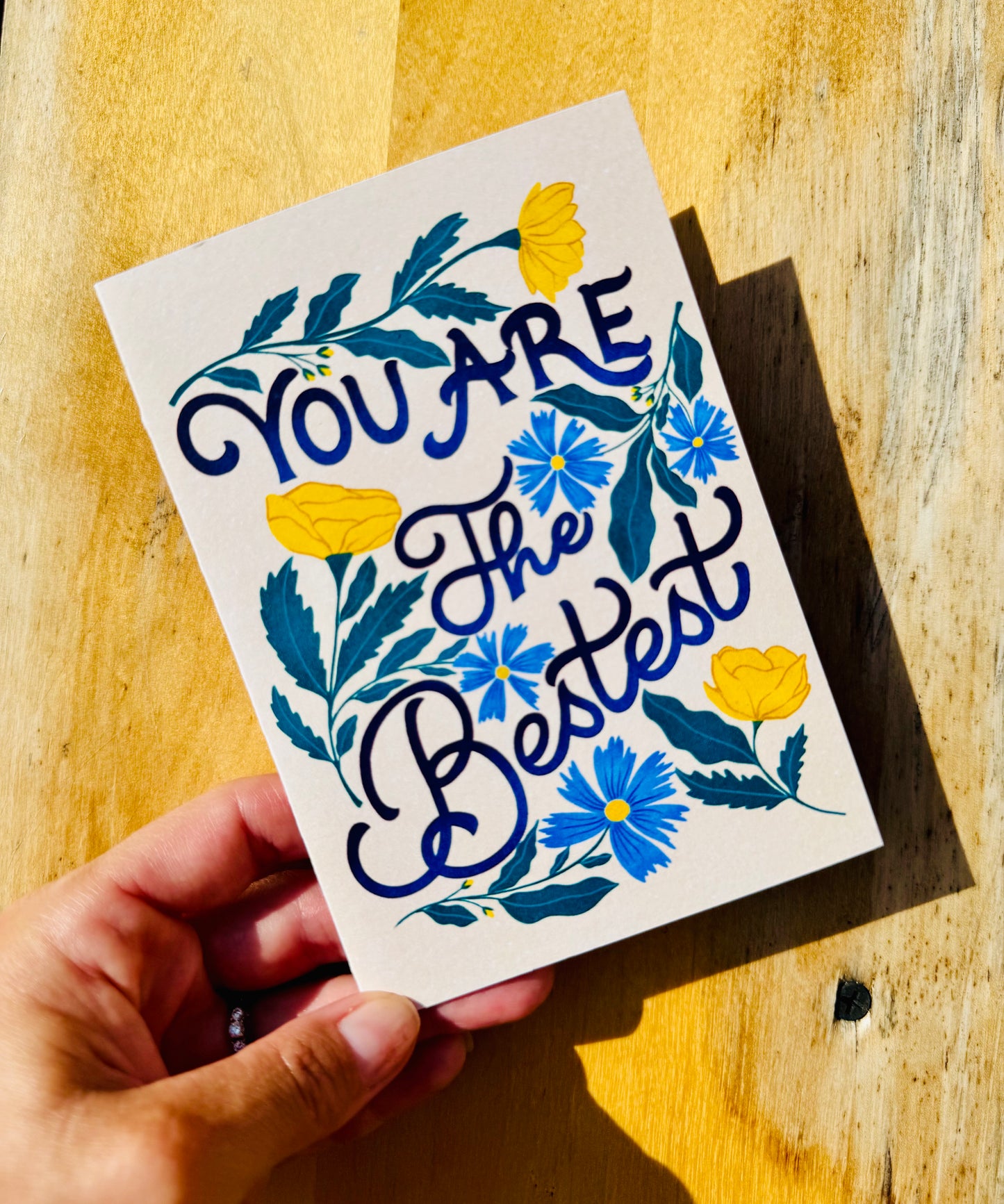 A hand holds a square white greeting card with the phrase 'YOU ARE The Bestest' in dark blue script, decorated with blue and yellow flowers and blue leaves, against a warm wooden background.