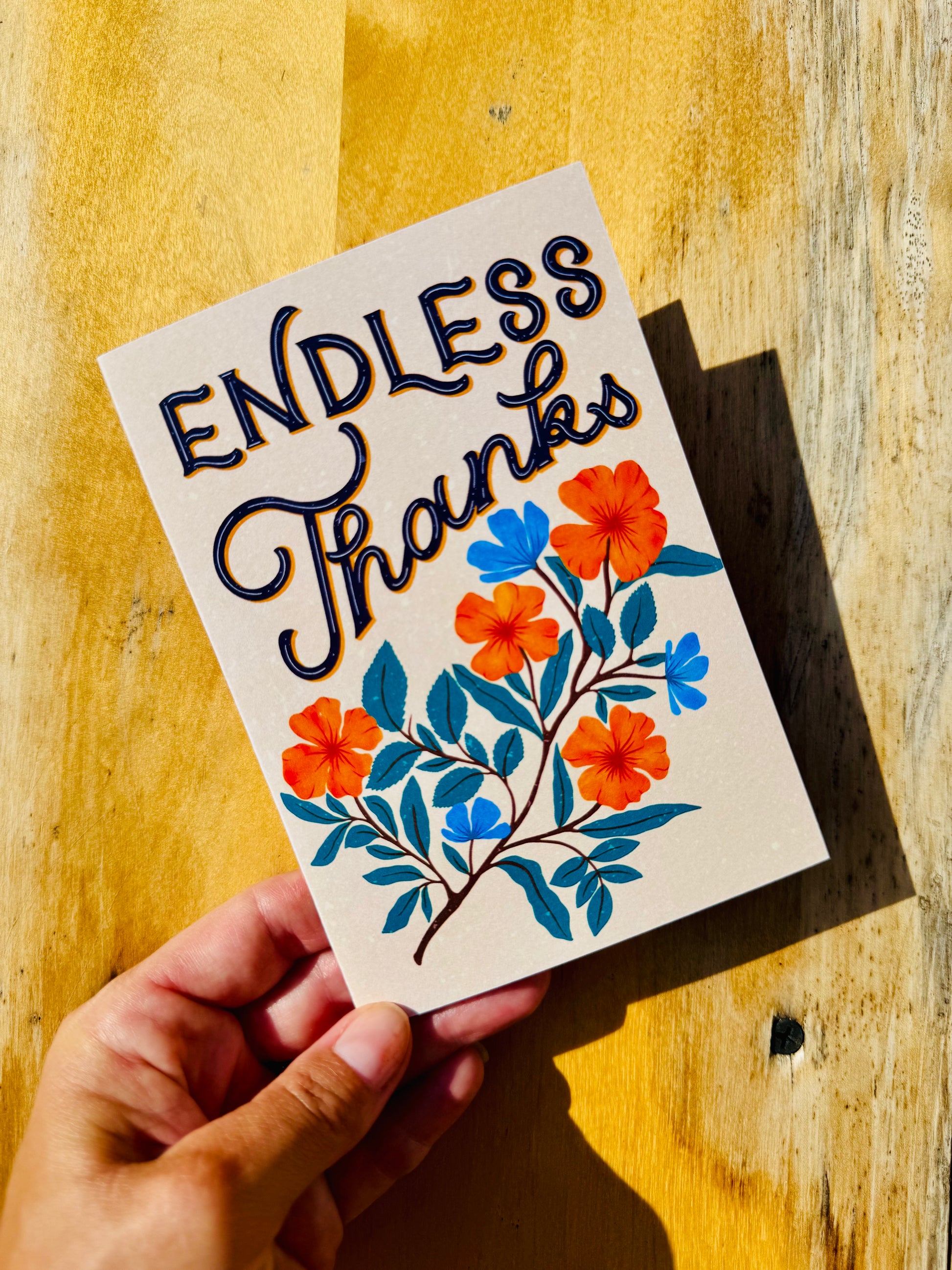 A hand holds a cream-colored greeting card that reads 'ENDLESS Thanks' in stylized black script with orange and blue floral illustrations, set against a warm yellow wooden background.