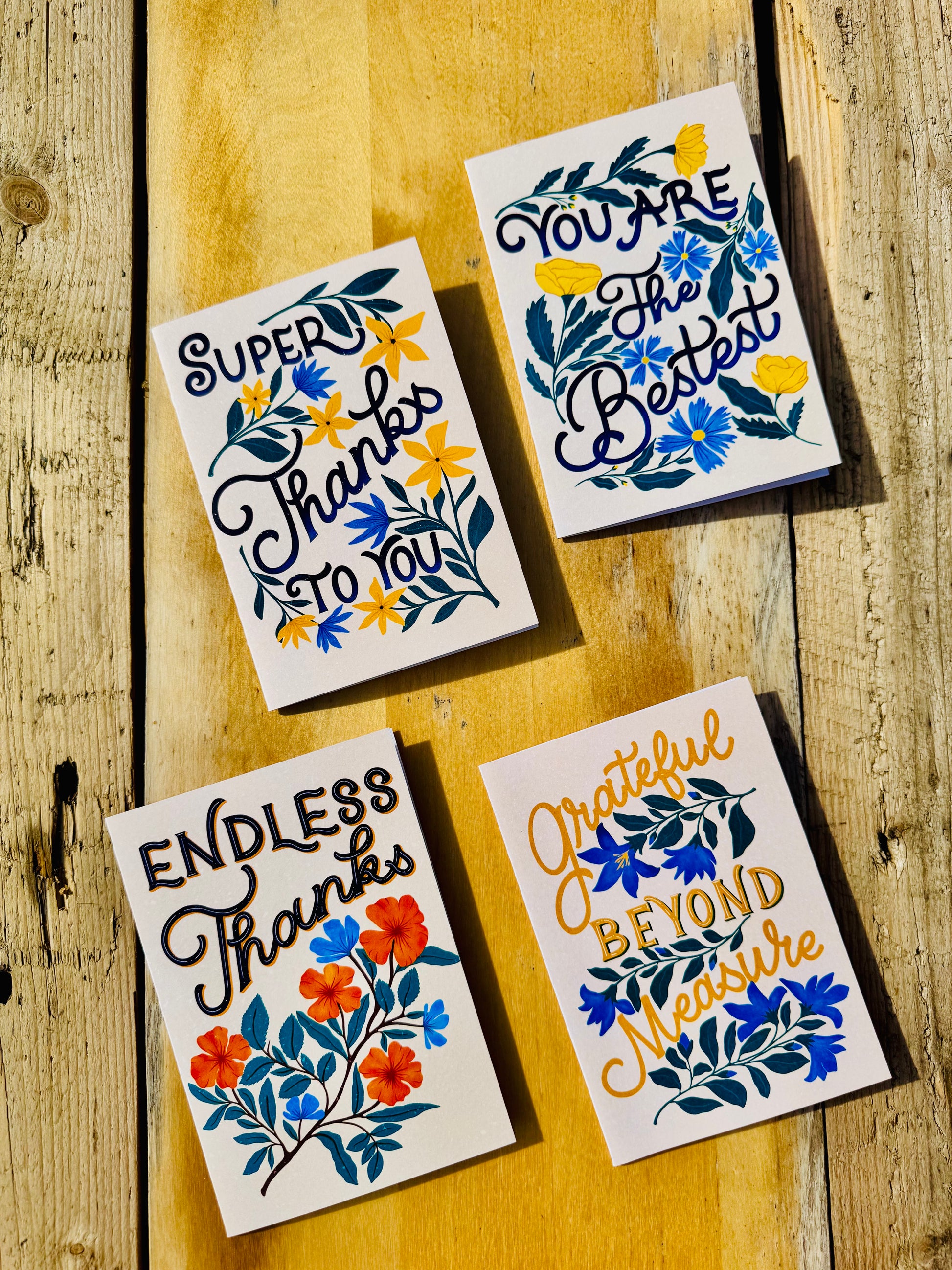 Four square thank-you cards with floral illustrations and inspirational thank-you messages, arranged on a rustic wooden surface.