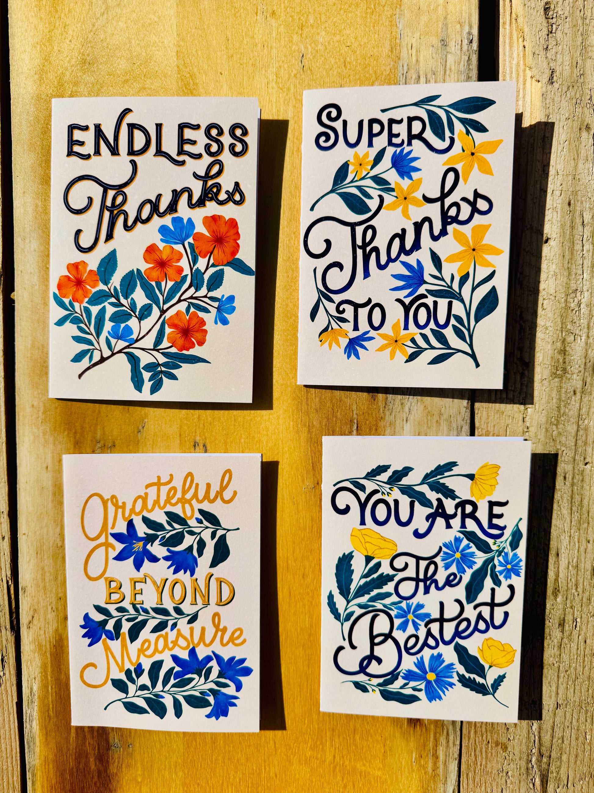 Four square thank-you cards with floral illustrations arranged on a rustic wooden surface. The cards feature different hand-lettered messages: 'ENDLESS Thanks', 'SUPER Thanks TO YOU', 'grateful BEYOND measure', and 'YOU ARE The Best'.