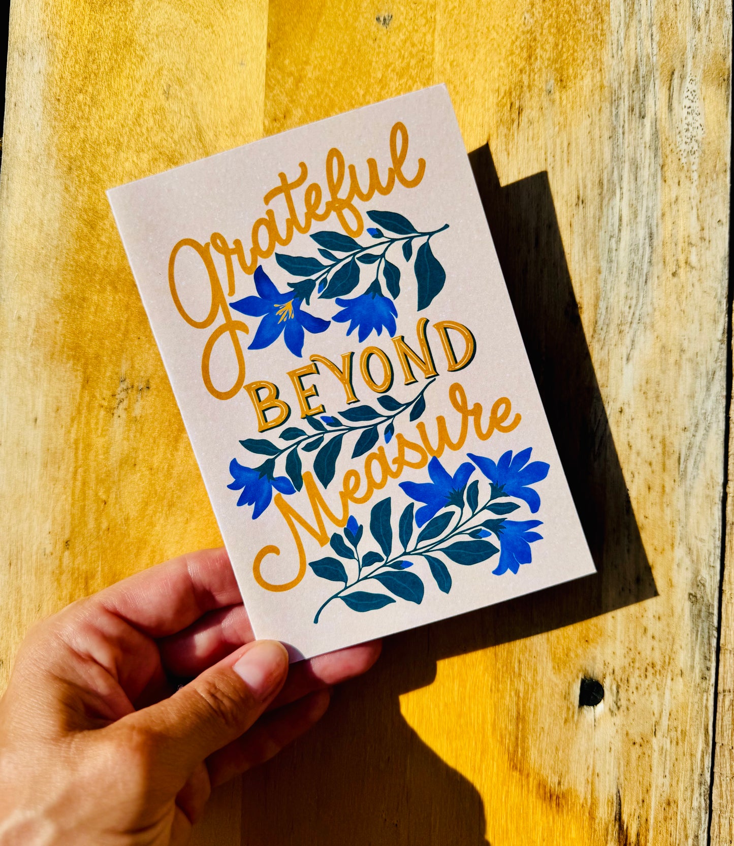 A hand holds a square art print or card with the phrase 'Grateful Beyond Measure' in gold script and block lettering, decorated with blue flowers and green leaves, against a rustic yellow wooden background.