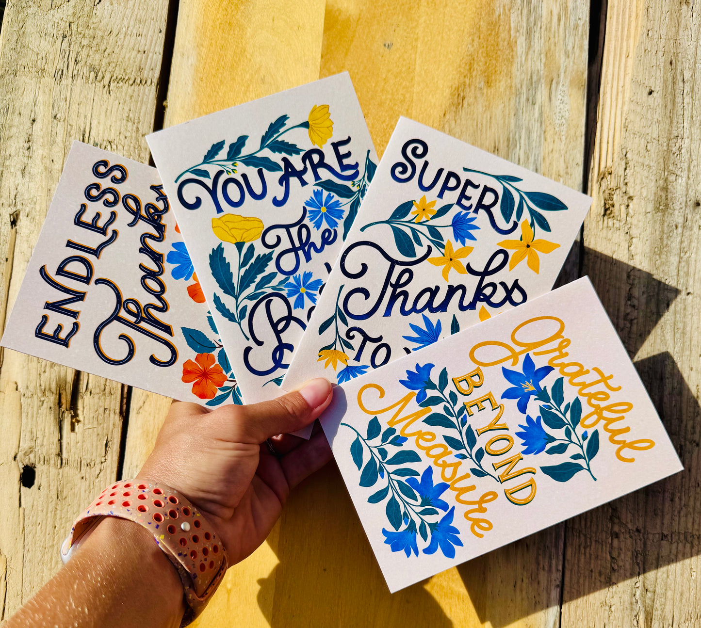 A hand holds four white greeting cards with colorful floral designs and inspirational text, photographed against a rustic wooden plank background.