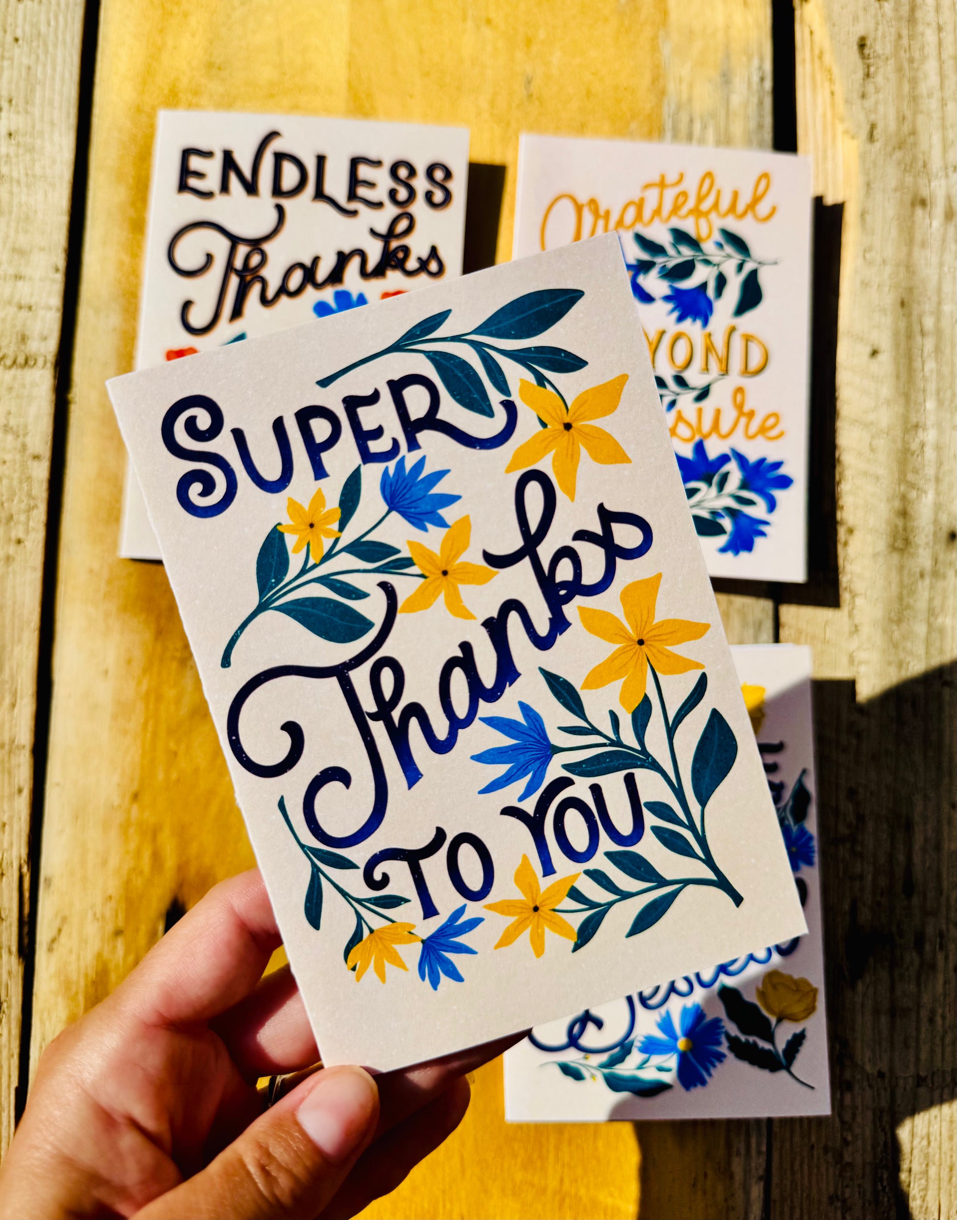 A hand holds a cream-colored thank you card with blue and yellow floral illustrations and the text 'SUPER Thanks to You' in dark blue script. Other thank you cards are stacked behind it on a rustic wooden surface.