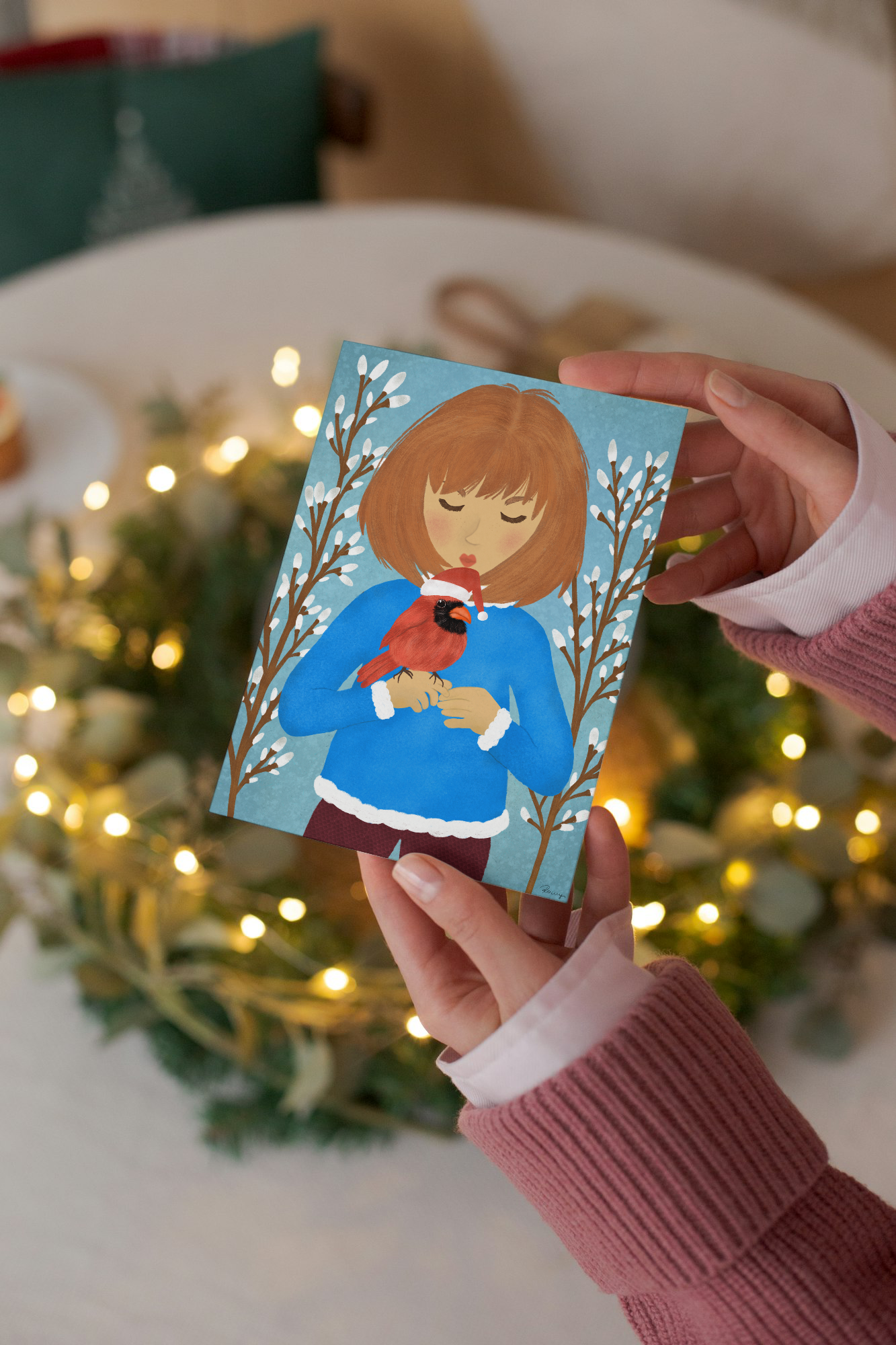 Person holding a Christmas card with a illustration of a person holding a cardinal bird, with festive lights and decorations in the background.