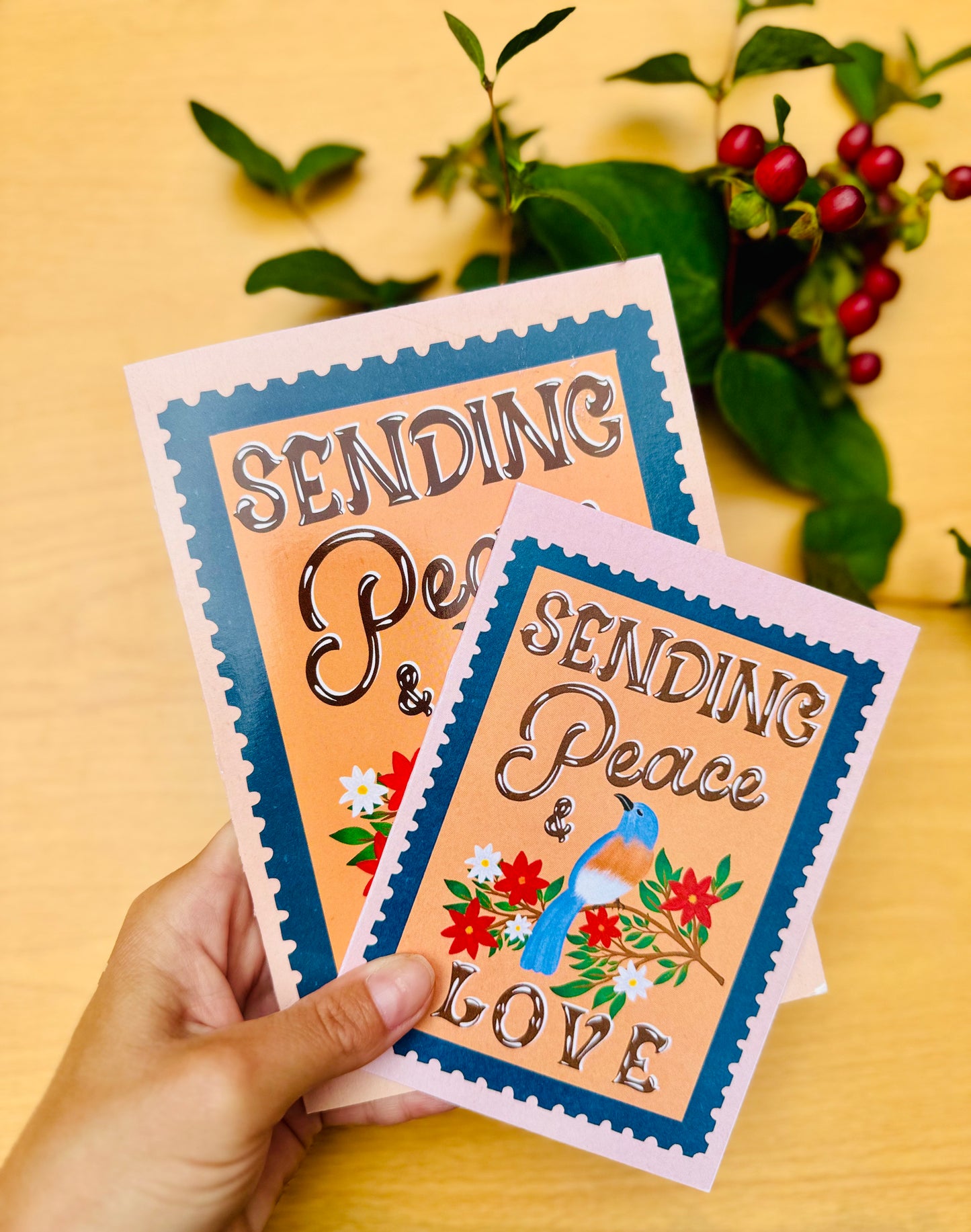 Two greeting cards with 'Sending Peace & Love' text held by a hand on a wooden surface with plants in the background.